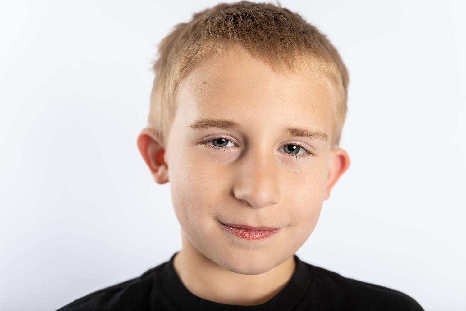 Young boy with blonde hair in black shirt smiling at camera against white background.