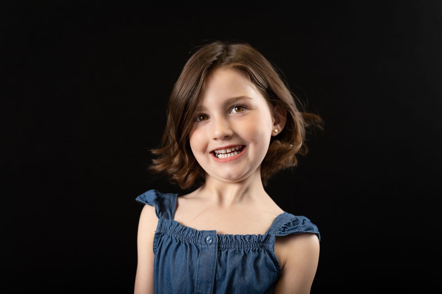 Happy young girl in a blue denim dress laughing against a black background.