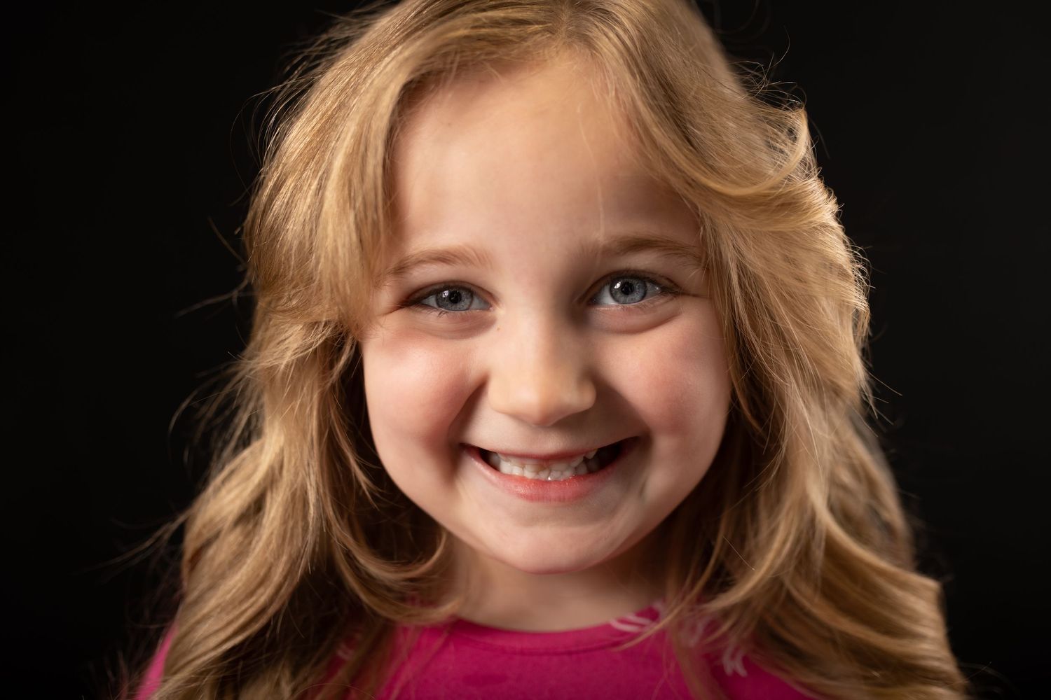 Young girl with long blonde hair smiling brightly wearing a pink top against a dark background.