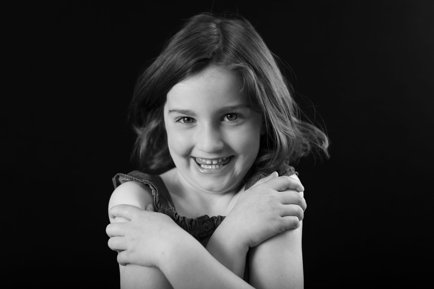 Black and white portrait of a smiling girl hugging herself against dark background.