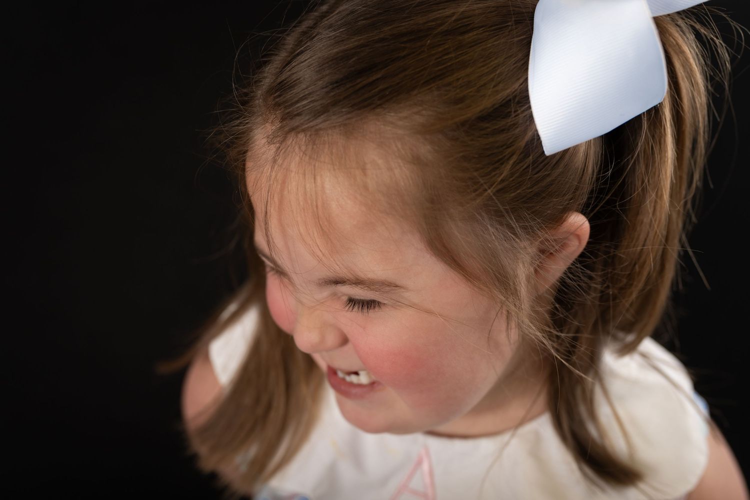 Young girl with a white bow in her hair smiling against a dark background.