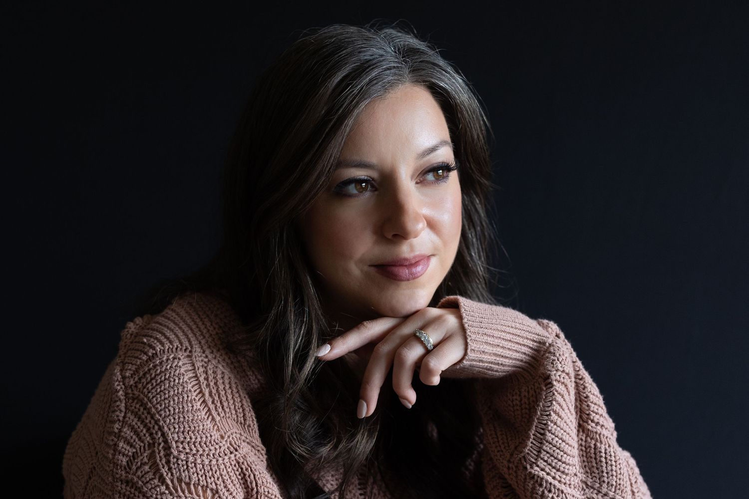 Woman with dark hair wearing a pink knit sweater posing against a dark background.