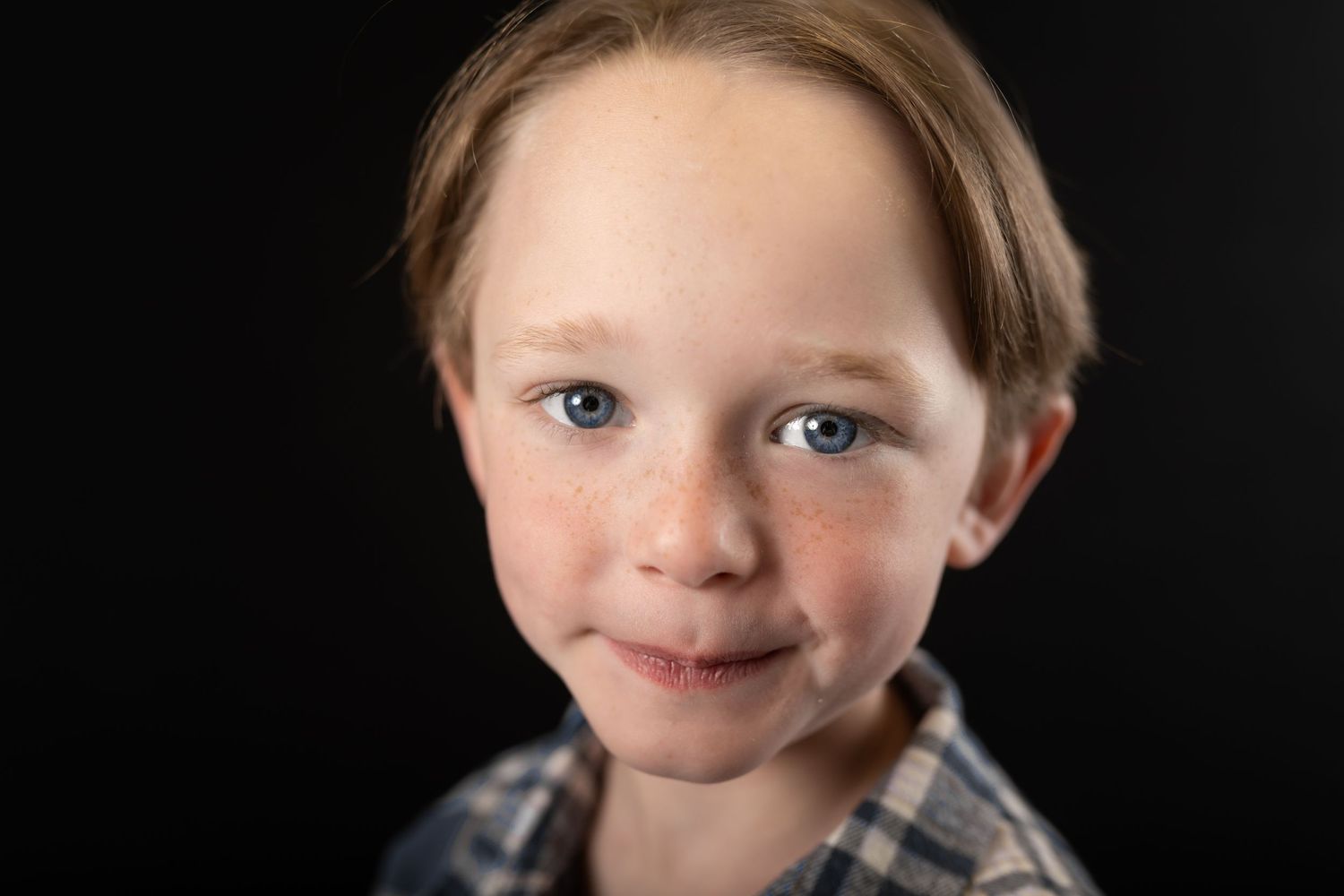 Freckled young boy with blue eyes and light brown hair smiling softly at camera against black background.