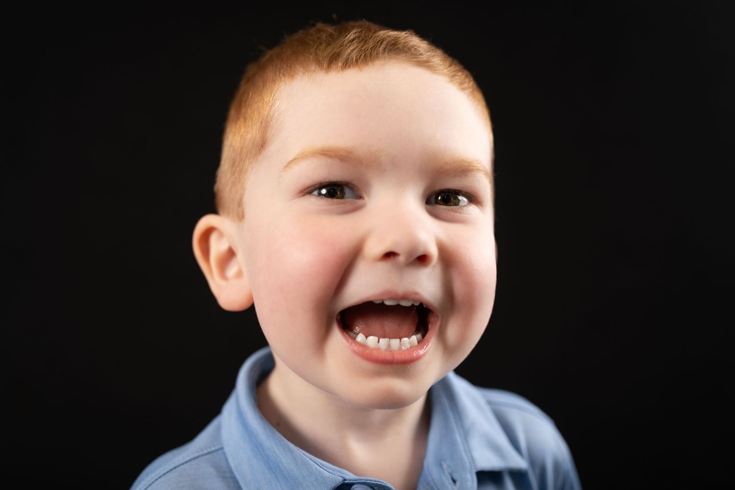 Excited young red-haired boy with mouth open wide laughing in a blue shirt, dark background.