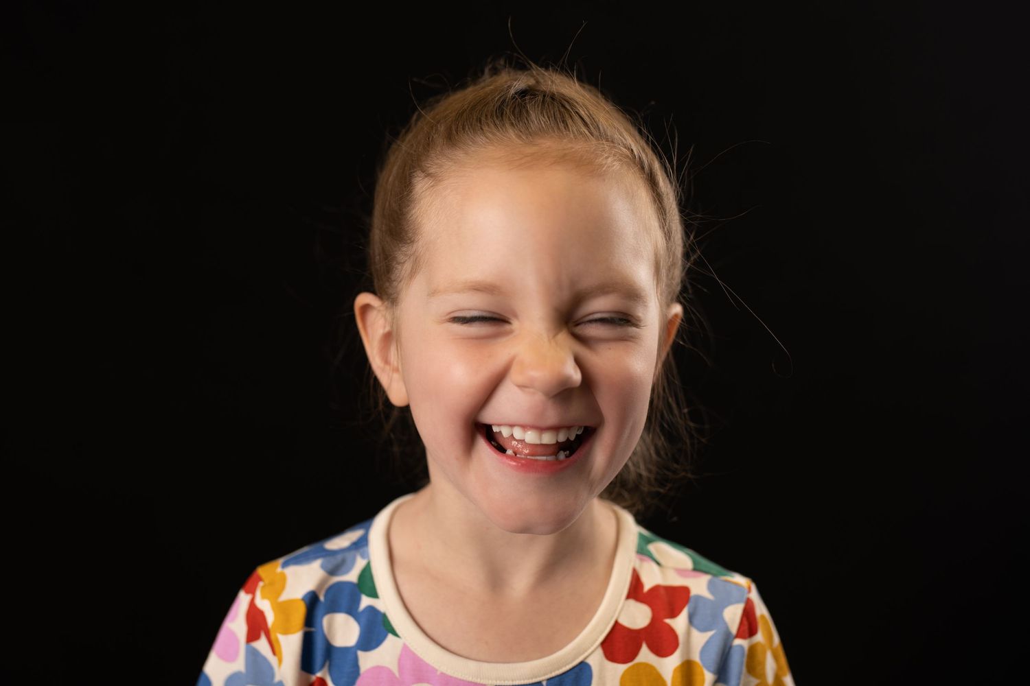 Laughing young girl in colorful floral shirt with eyes closed against black background.