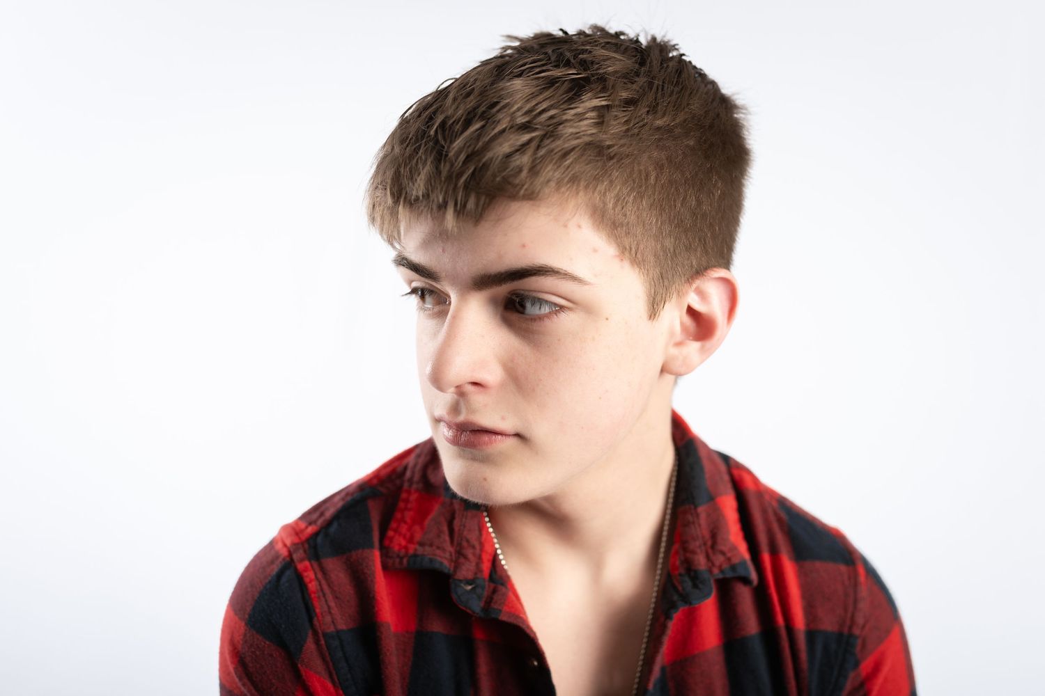 Teenage boy in red plaid shirt looking sideways against white background.