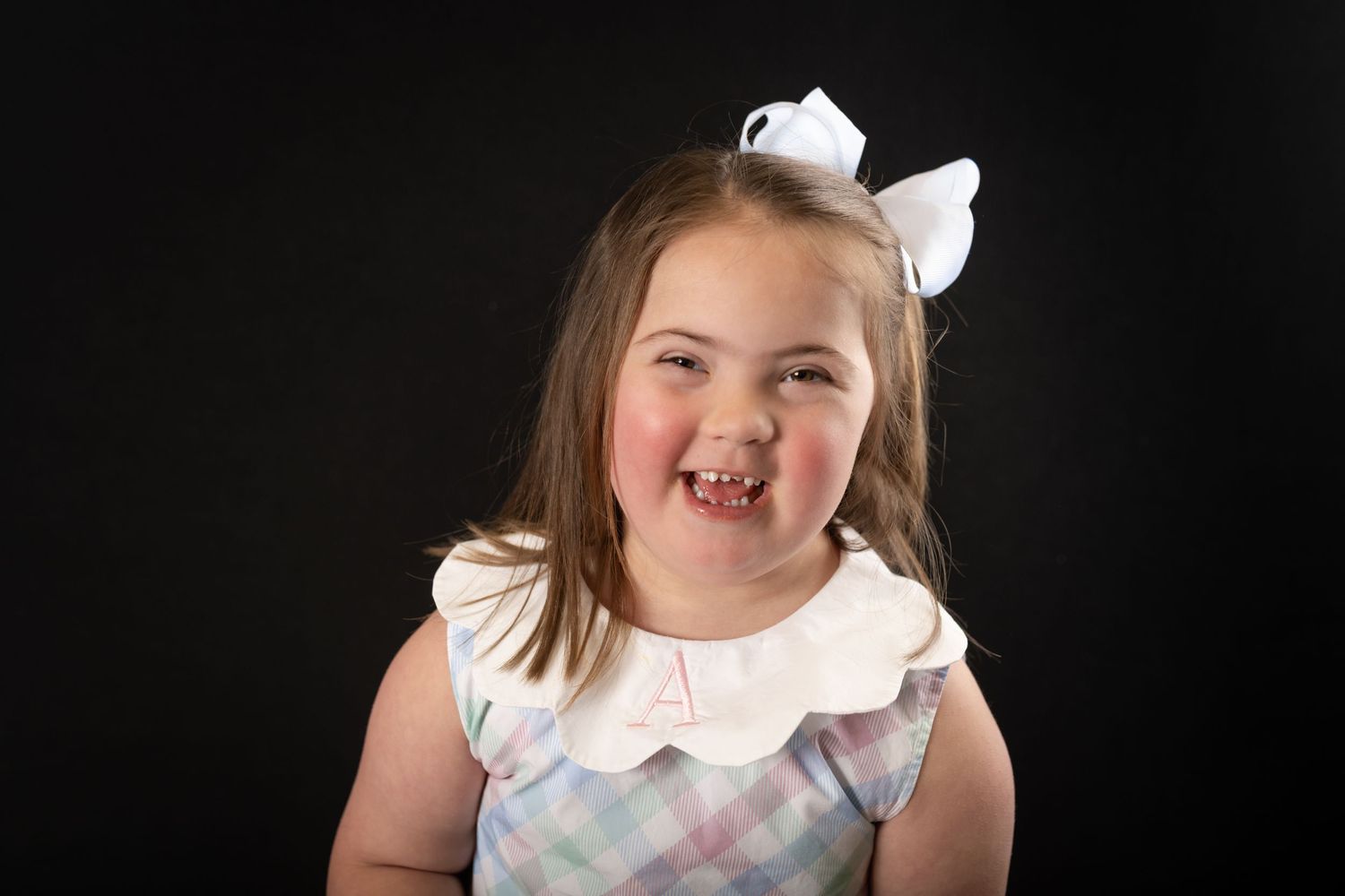 Joyful young girl with Down syndrome wearing white bow and plaid dress against black background.