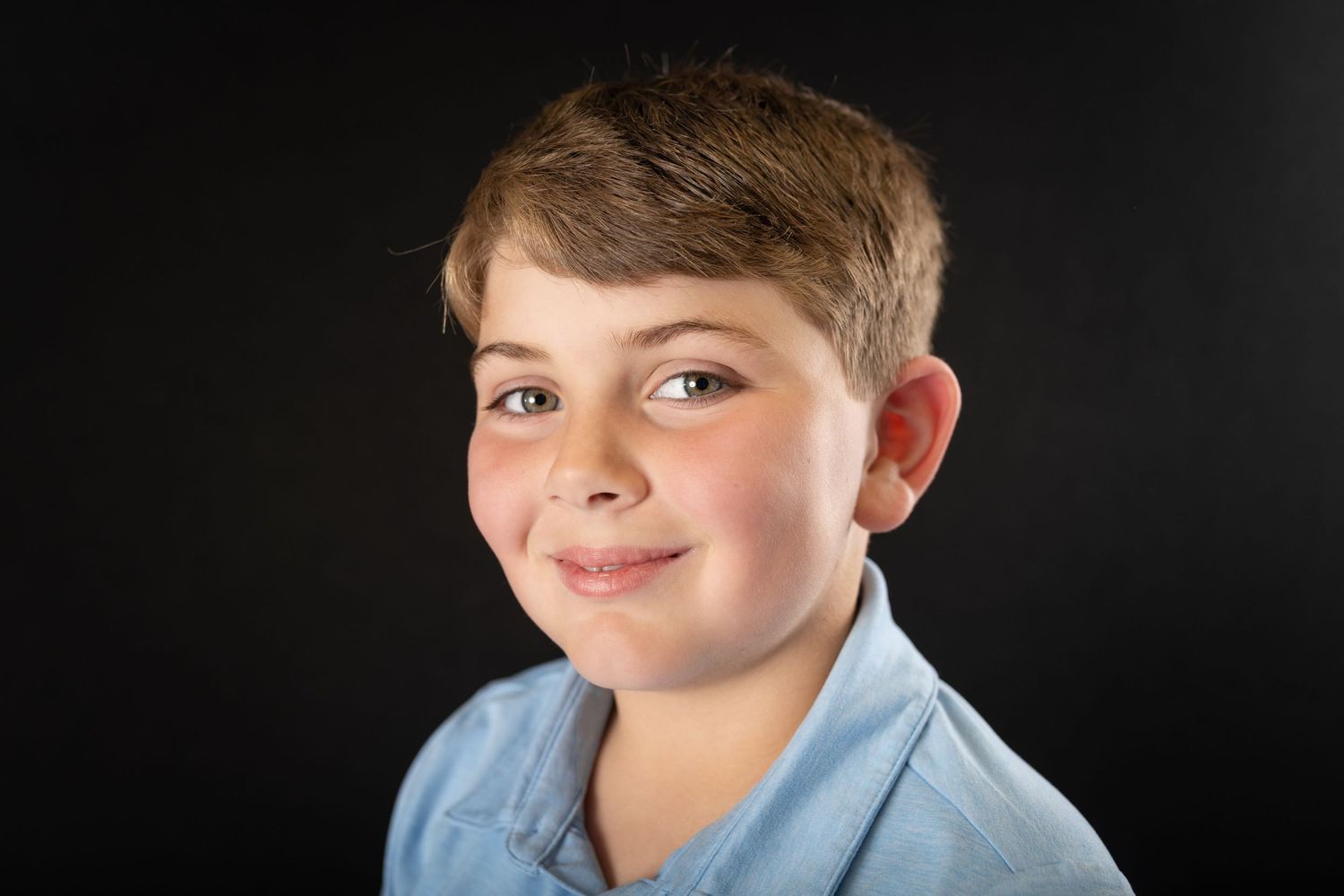 Smiling young boy with short brown hair wearing a light blue shirt against a dark background.