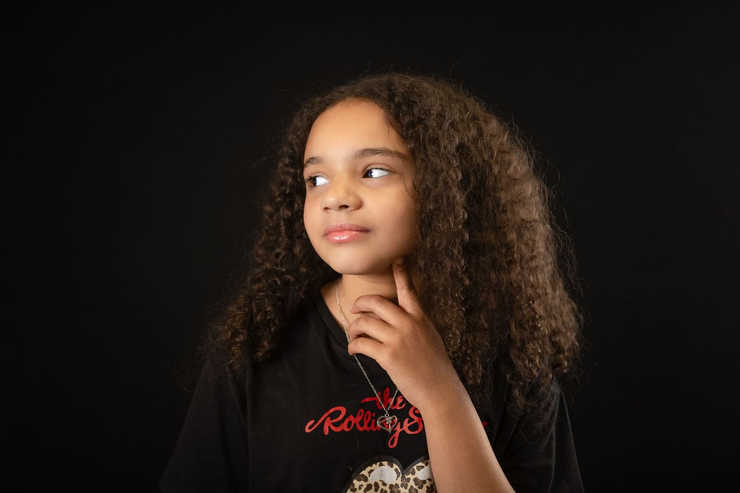 Young girl with long curly hair posing thoughtfully against a dark background in a black jacket.