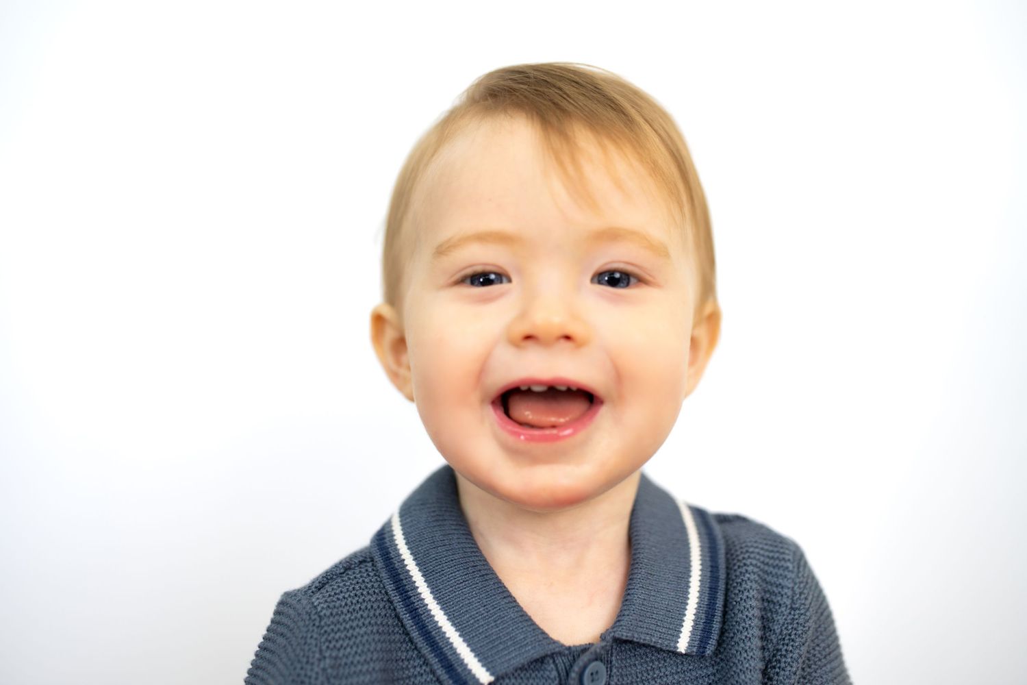 Smiling toddler boy in grey polo shirt with blonde hair against white background.