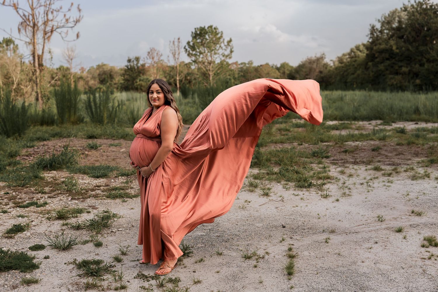Pregnant woman in flowing coral dress posing outdoors with fabric blowing in wind.