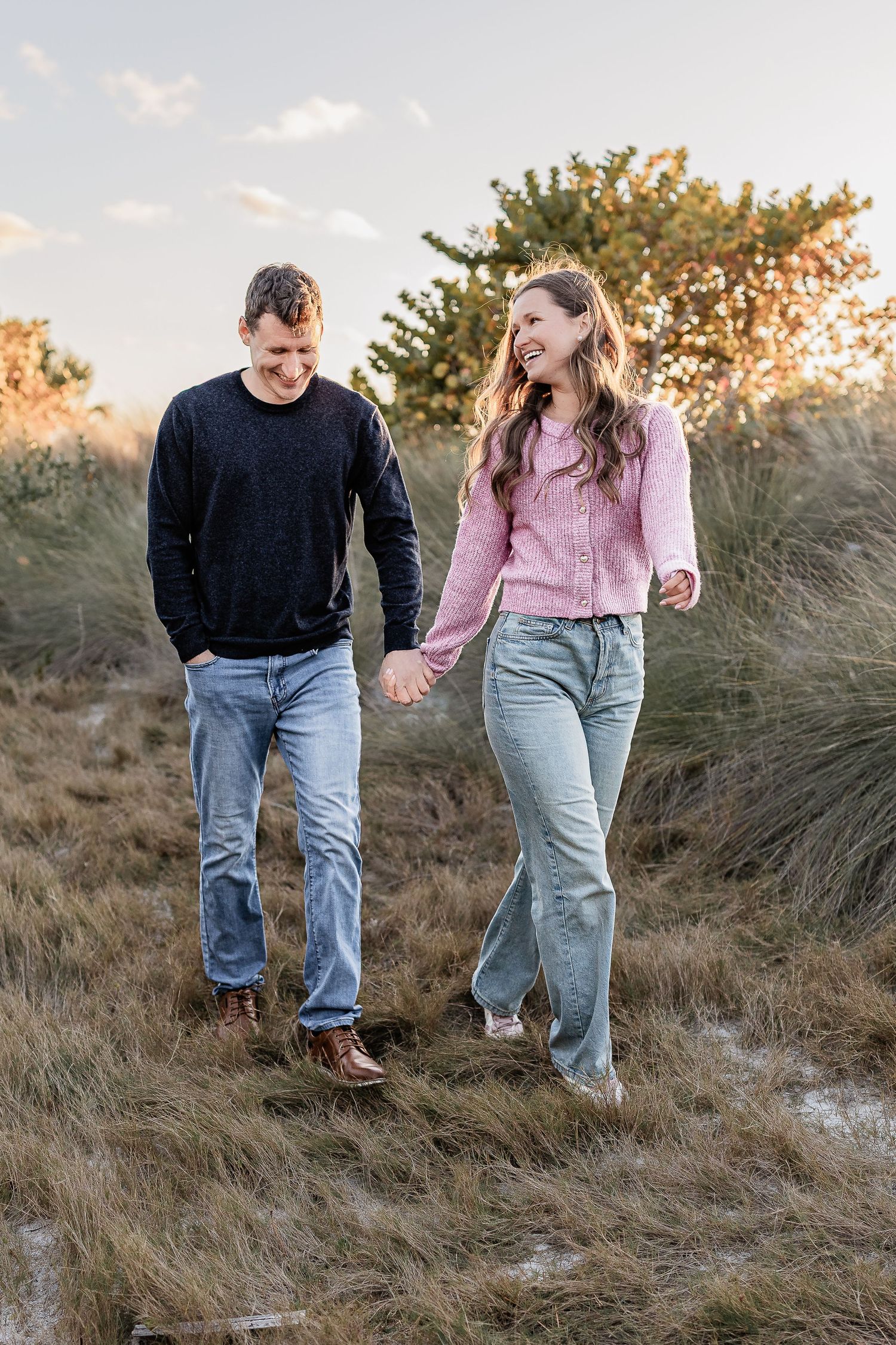 Young couple holding hands and walking through golden coastal grass at sunset.