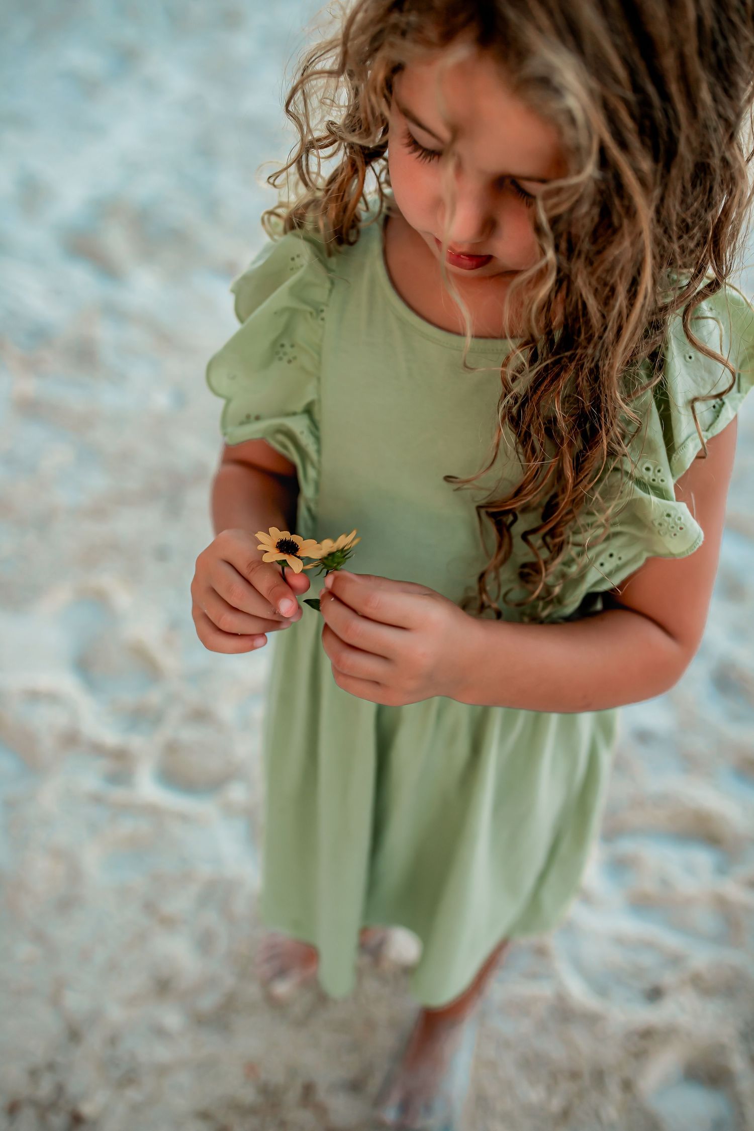 A young girl with curly hair in a green dress holds a small flower at the beach.