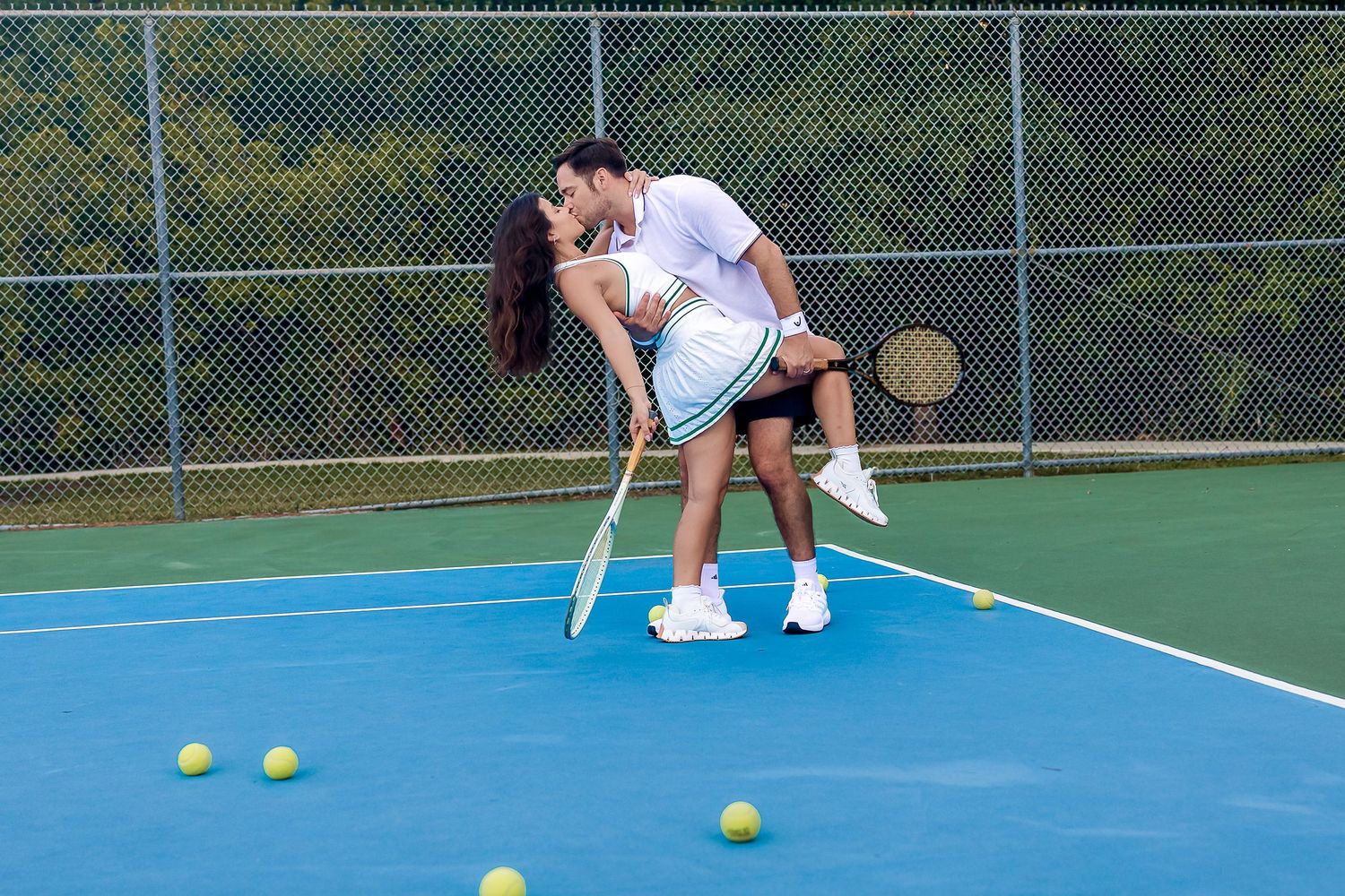 Couple sharing a romantic kiss on a blue tennis court surrounded by tennis balls and rackets.