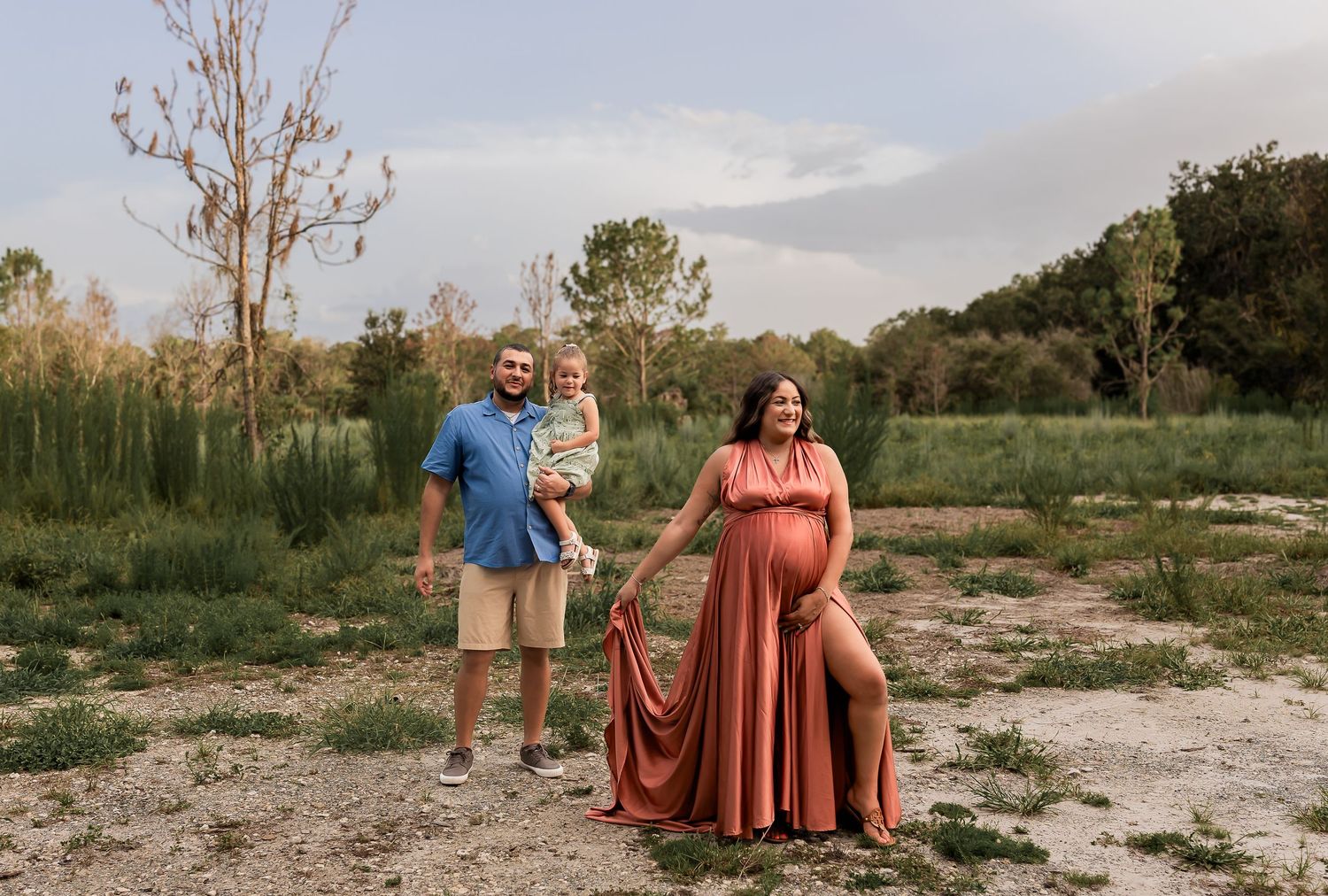 Expecting couple with toddler poses outdoors at sunset, woman wearing elegant rust maternity gown.