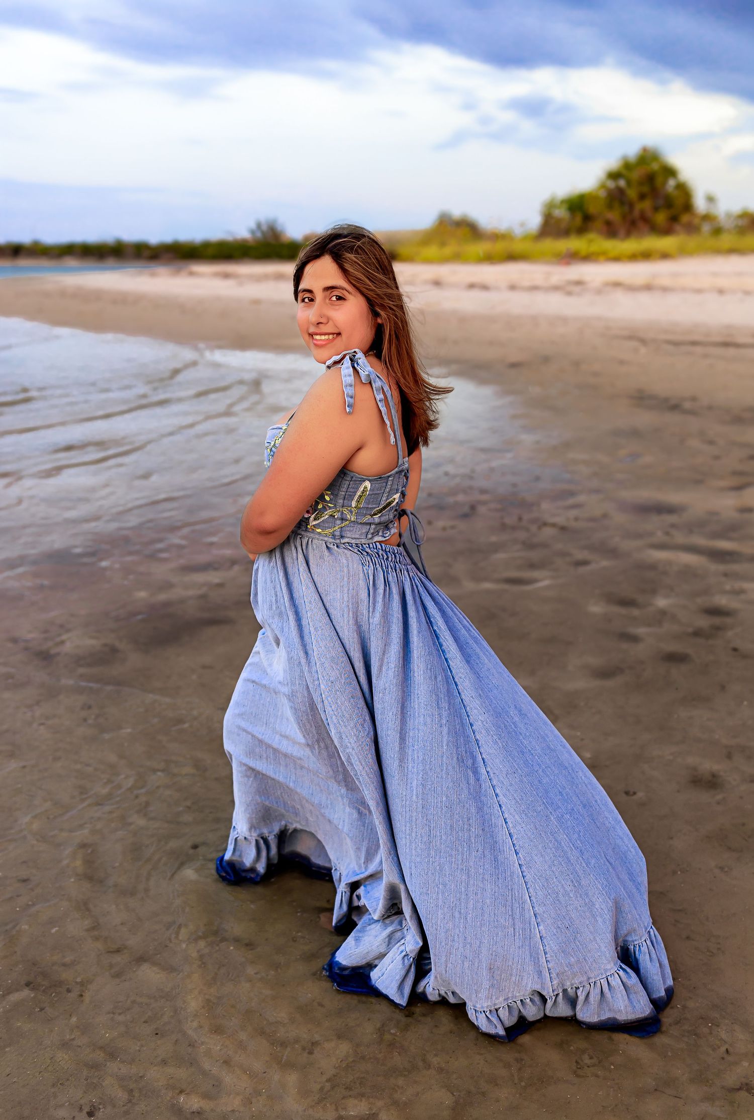 Young woman in flowing blue halter dress posing on a sandy beach with ocean waves behind her.
