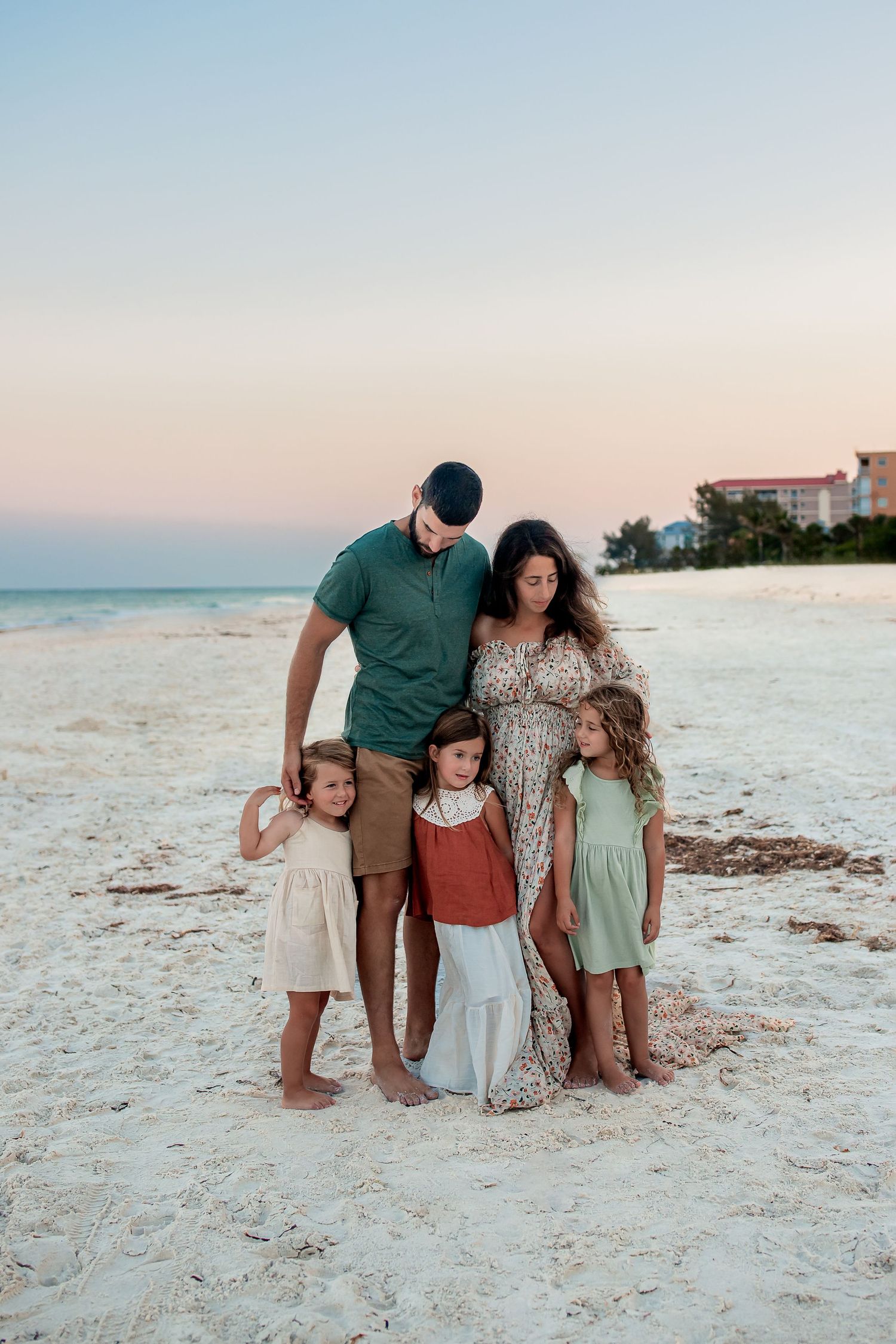 Family of five poses on white sand beach at sunset, parents embracing three young daughters in colorful dresses.