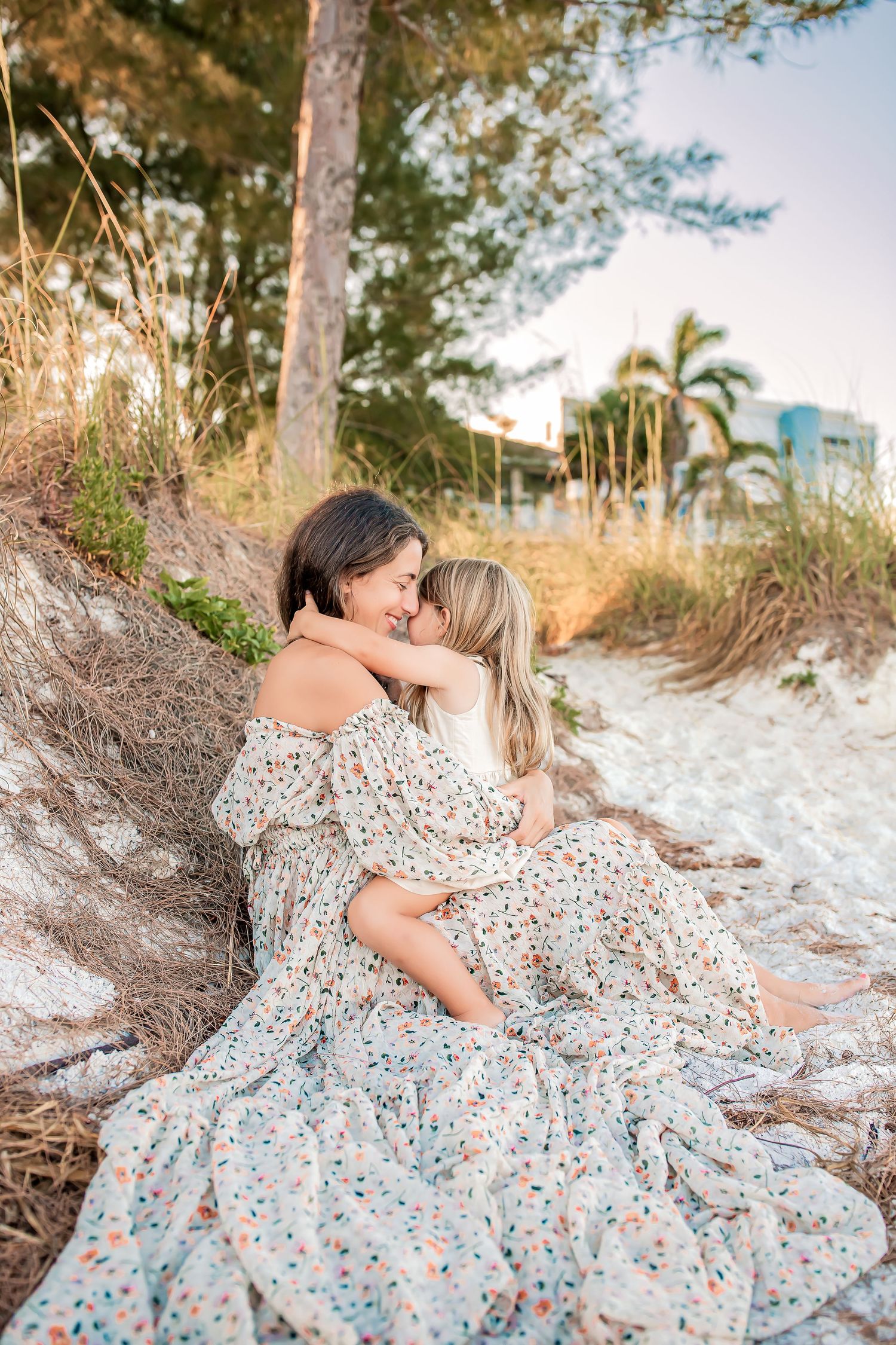 Mother and daughter sharing tender embrace while sitting against tree roots on sandy beach at golden hour.