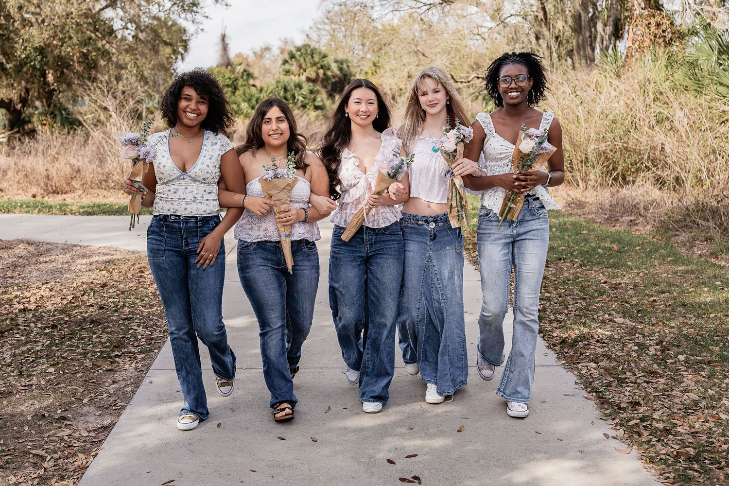 Five diverse young women laugh and walk together on a path carrying flowers in a park.