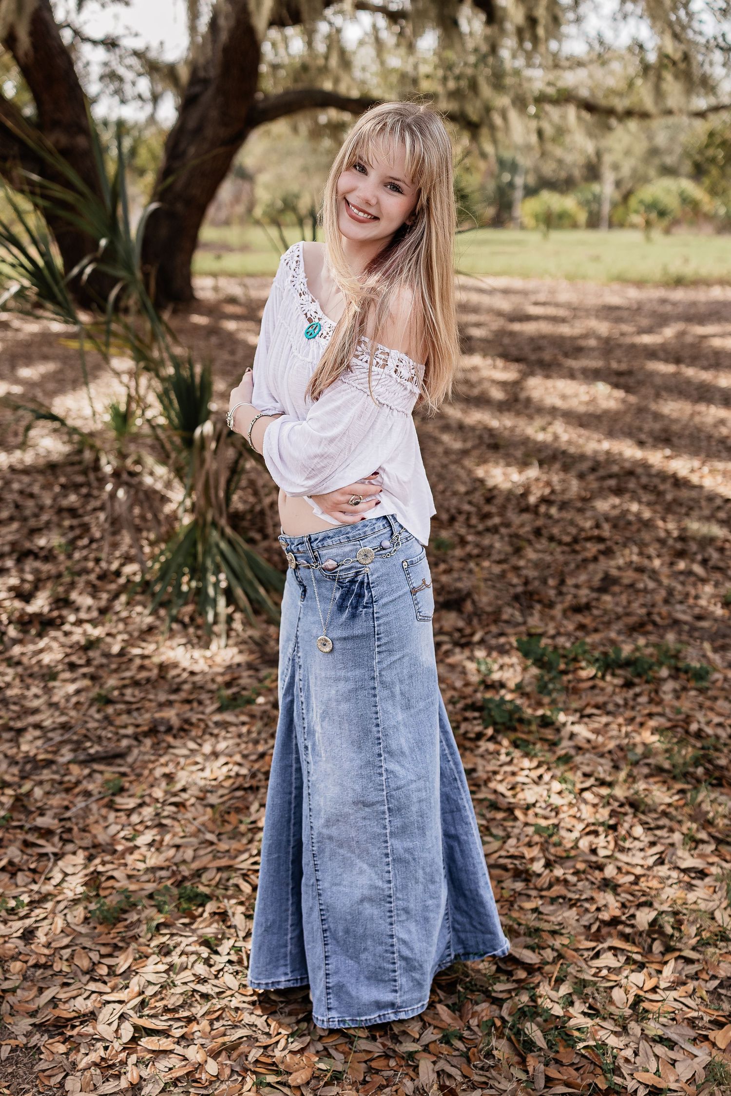 Blonde teen girl smiles wearing white boho top and long denim skirt among fallen leaves under oak tree.