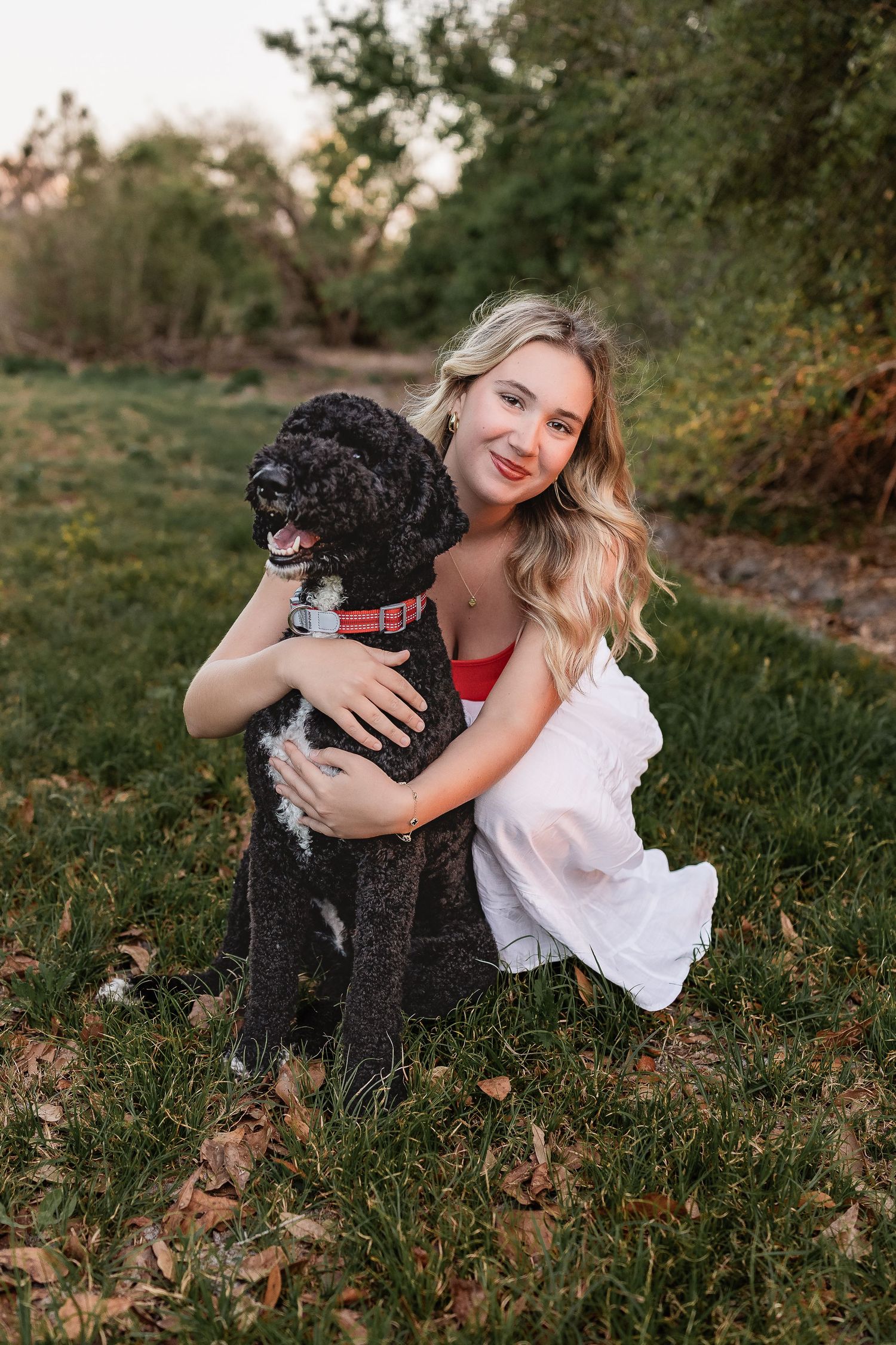Blonde girl in white dress hugging a black curly-haired dog on a grassy lawn at sunset.