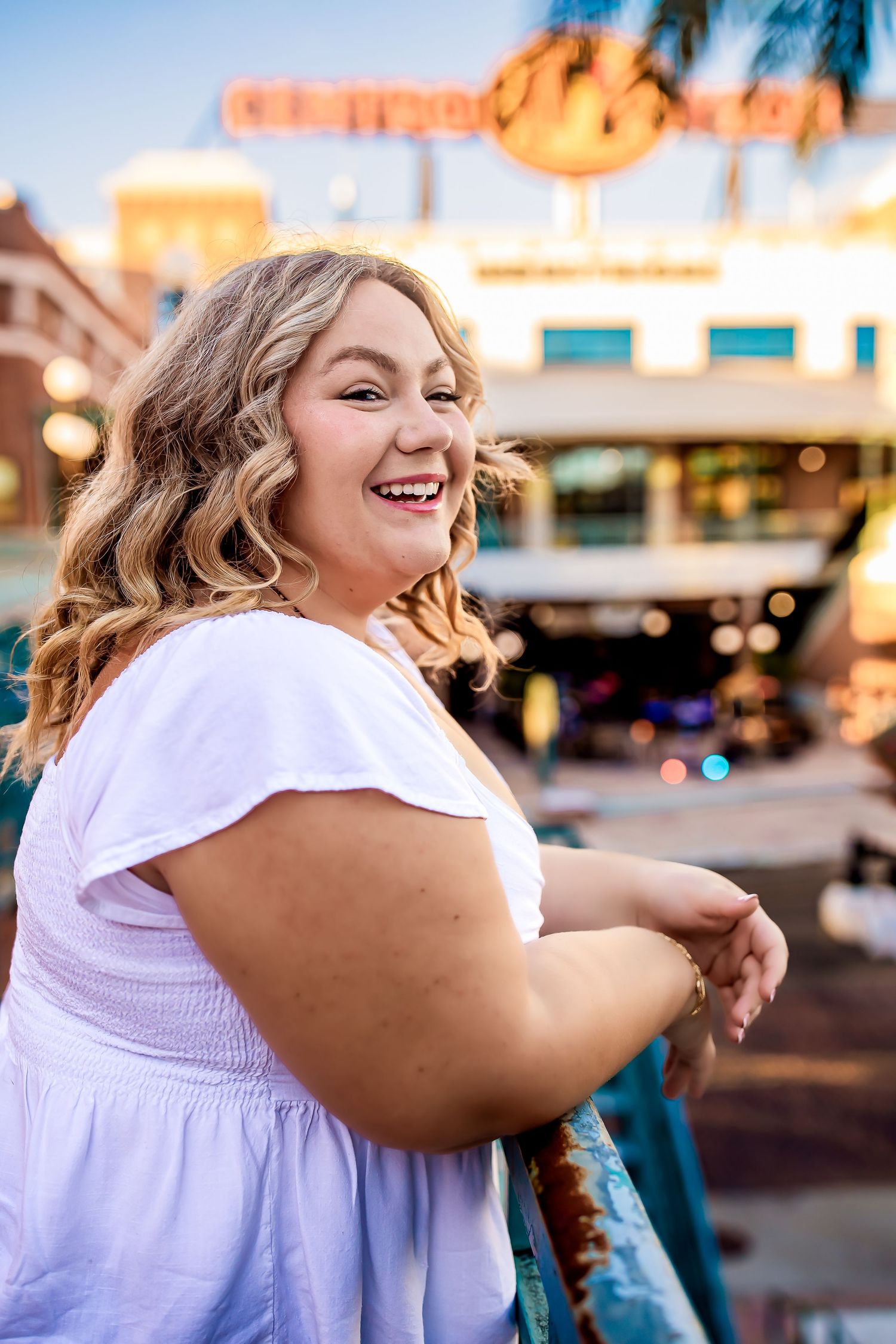 Happy blonde woman in white dress laughing while leaning on railing at an outdoor venue at dusk.