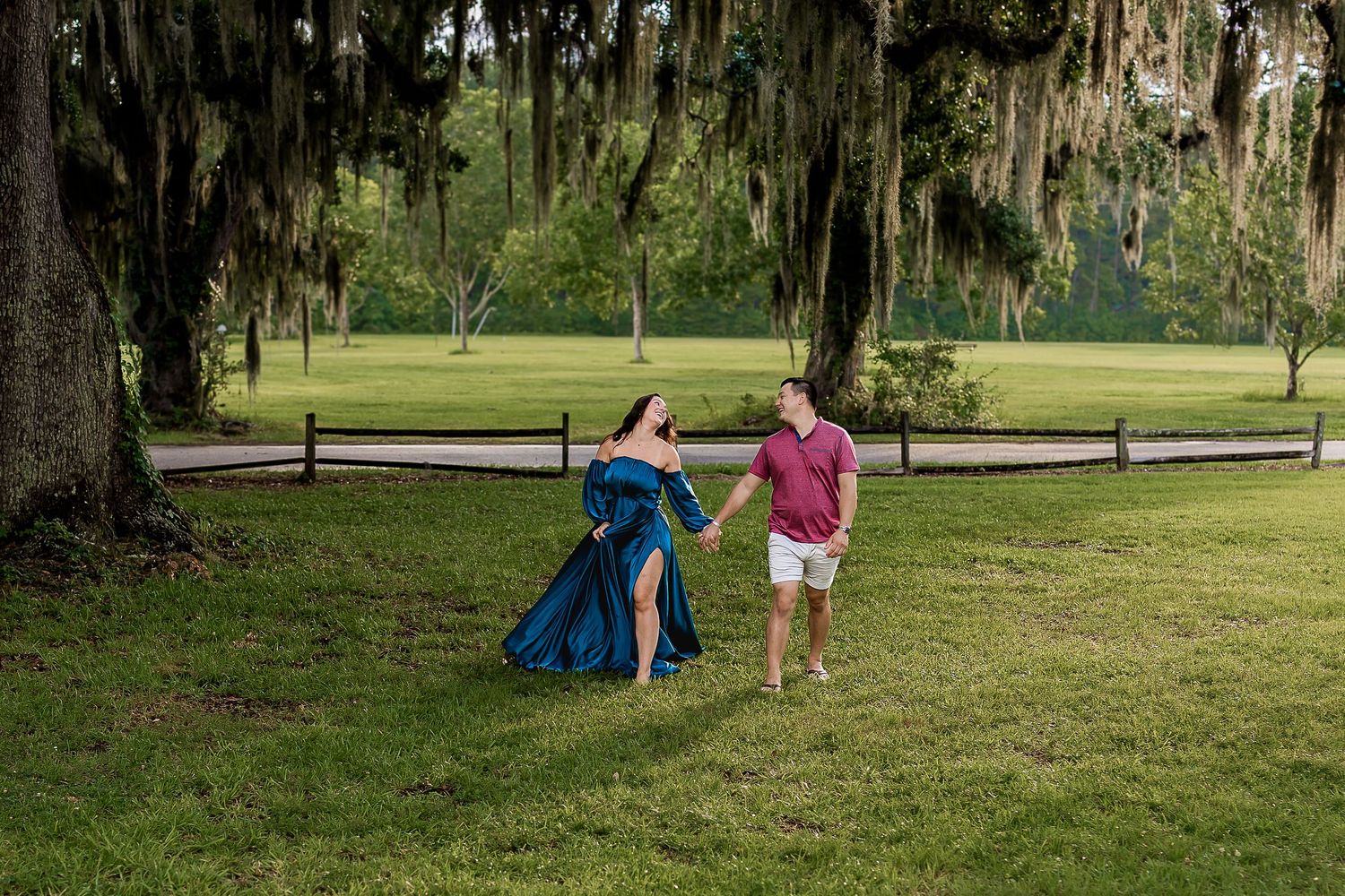 A pregnant woman in a flowing teal dress holds hands with her partner walking under moss-draped trees.