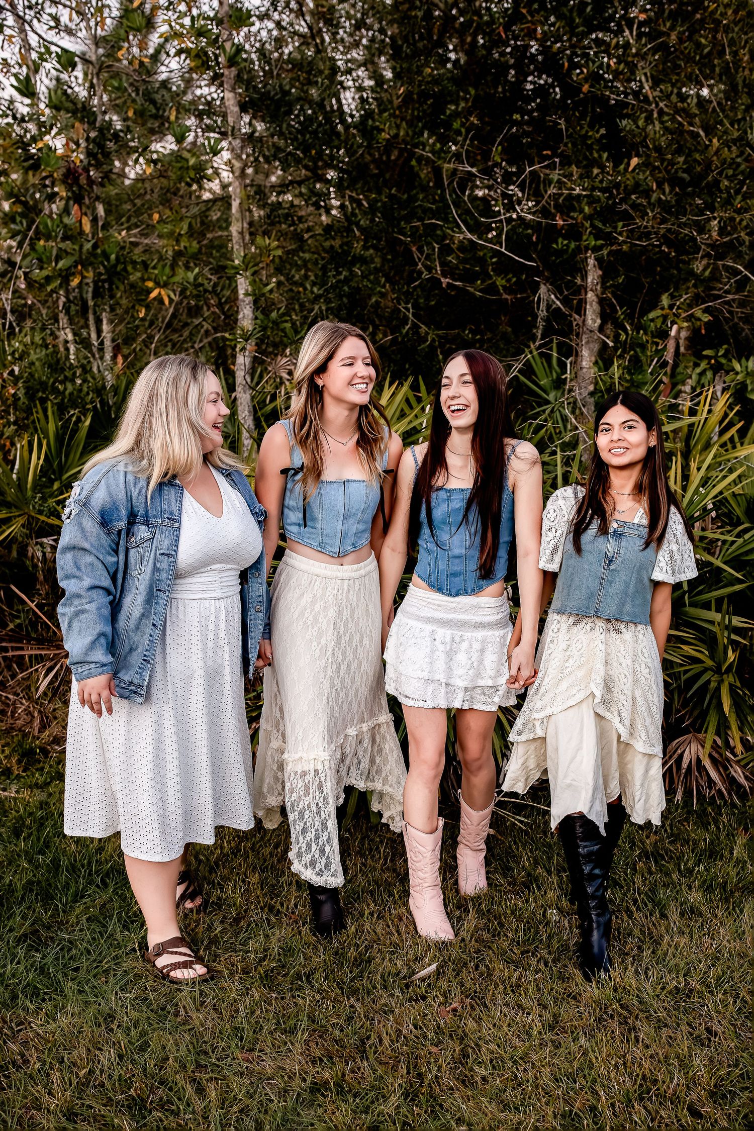 High school friends in white dresses and denim jackets laughing together outdoors among lush green foliage.