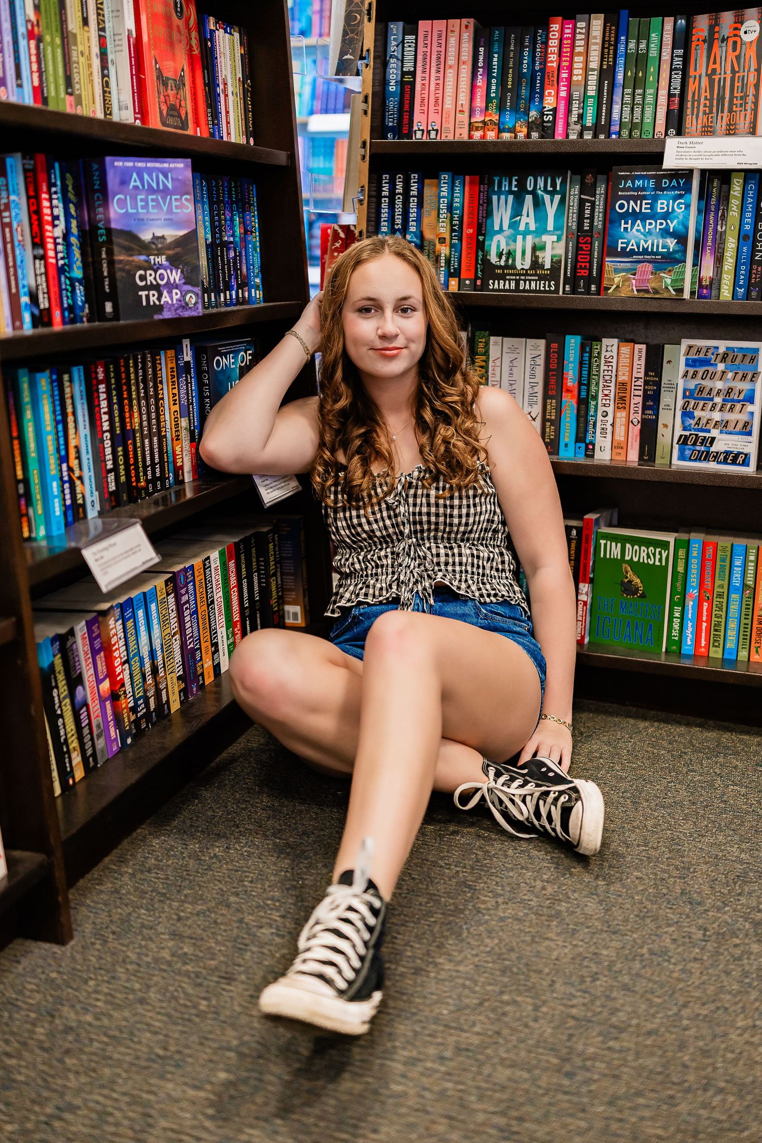 Young womanl in plaid top and denim shorts sitting on floor in front of bookstore shelves.
