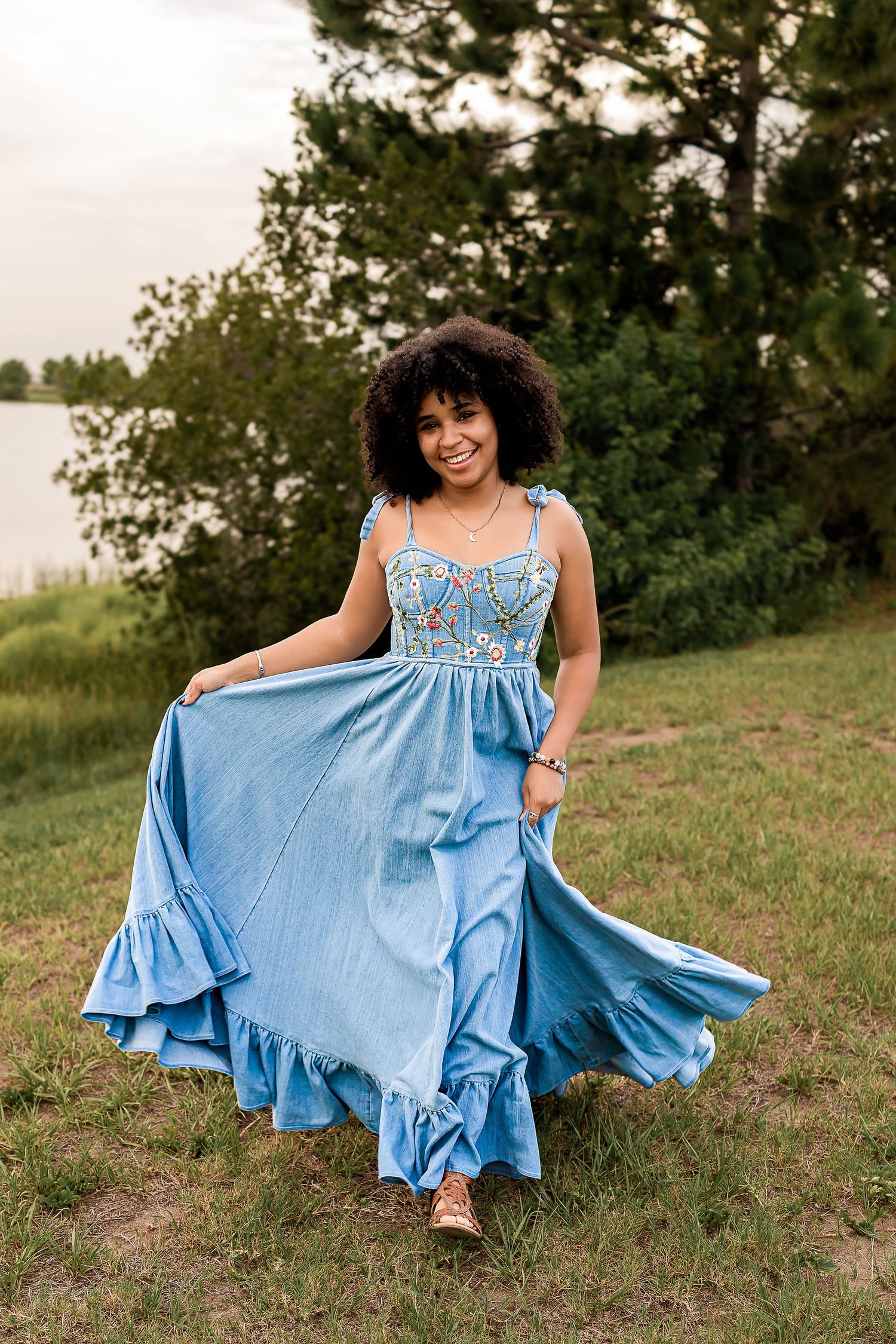 Young woman in blue floral maxi dress twirling outdoors near a lake surrounded by green trees.