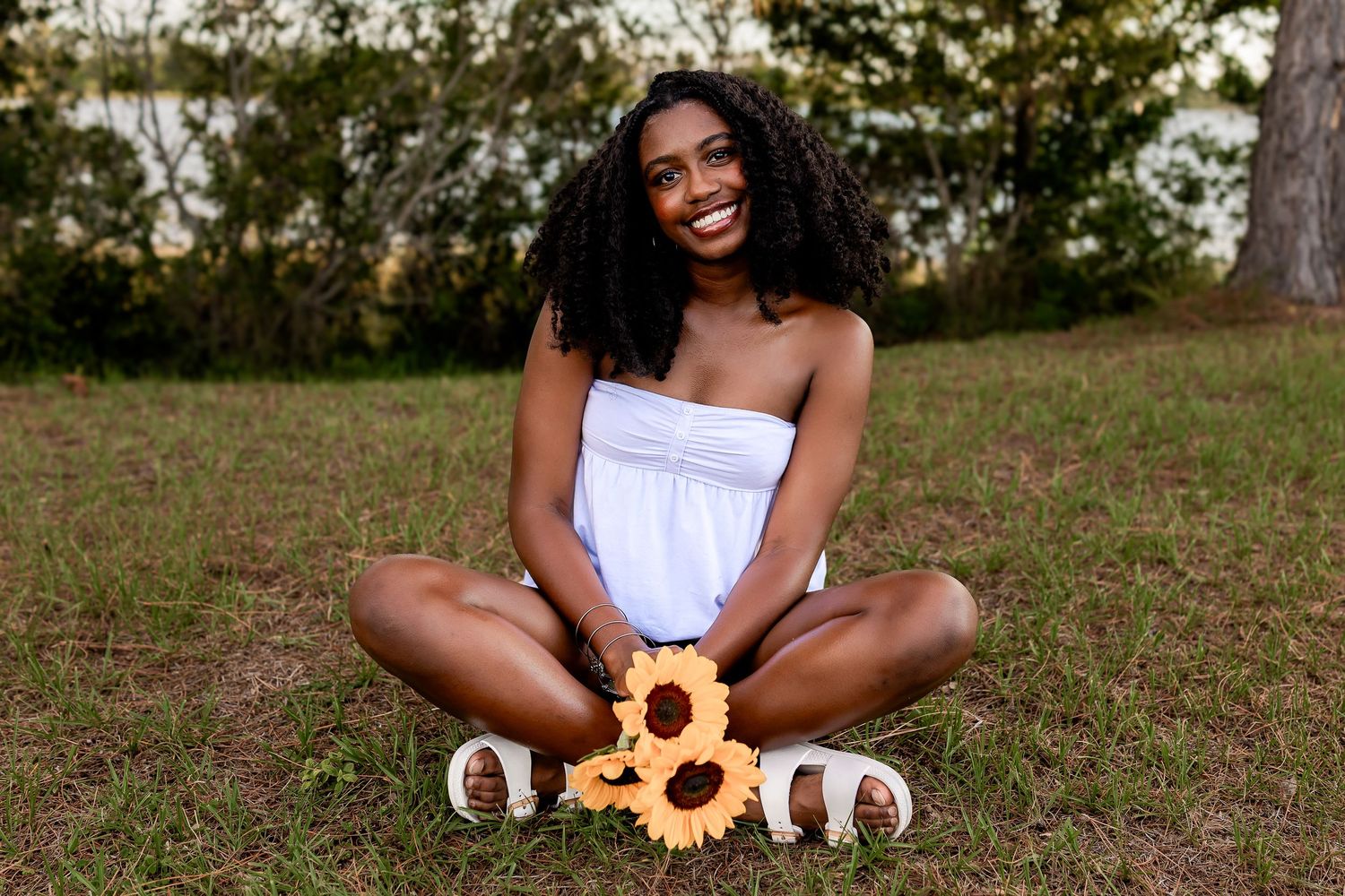 Young woman in white dress sitting cross-legged on grass holding sunflowers outdoors.