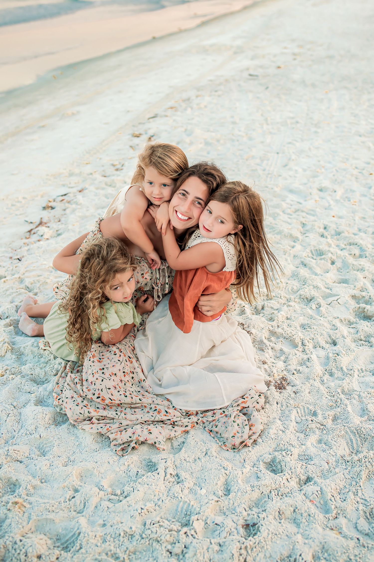 Mother sitting on beach sand hugging three young daughters warmly.