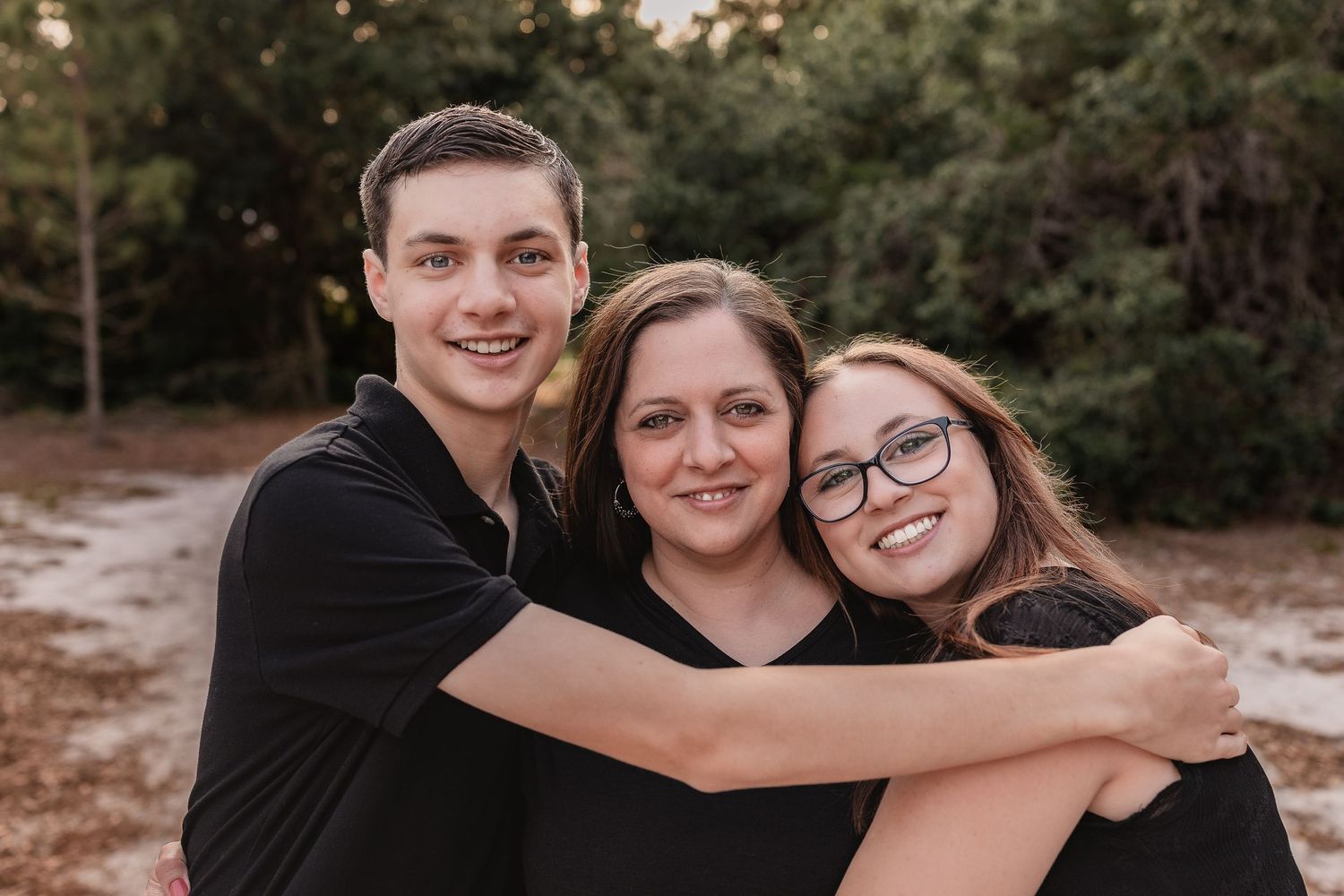 Three family members in black outfits smile and embrace outdoors in a wooded setting.
