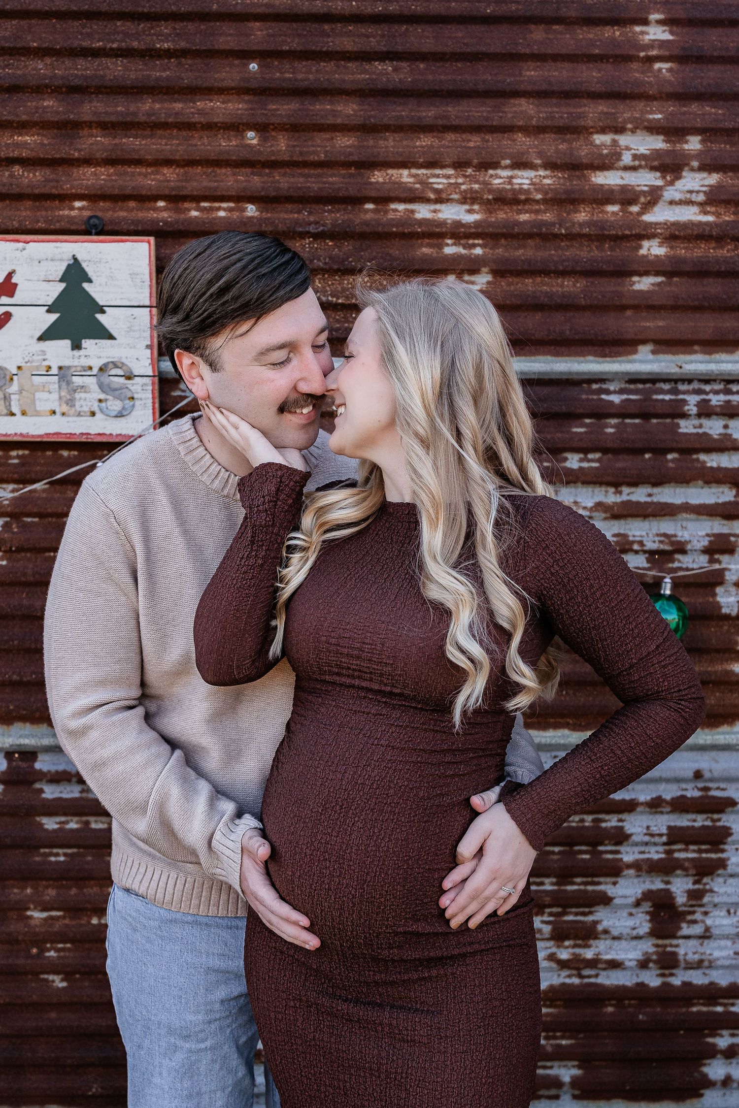 Expecting couple sharing a tender kiss outdoors against a rustic metal wall during maternity photo session.