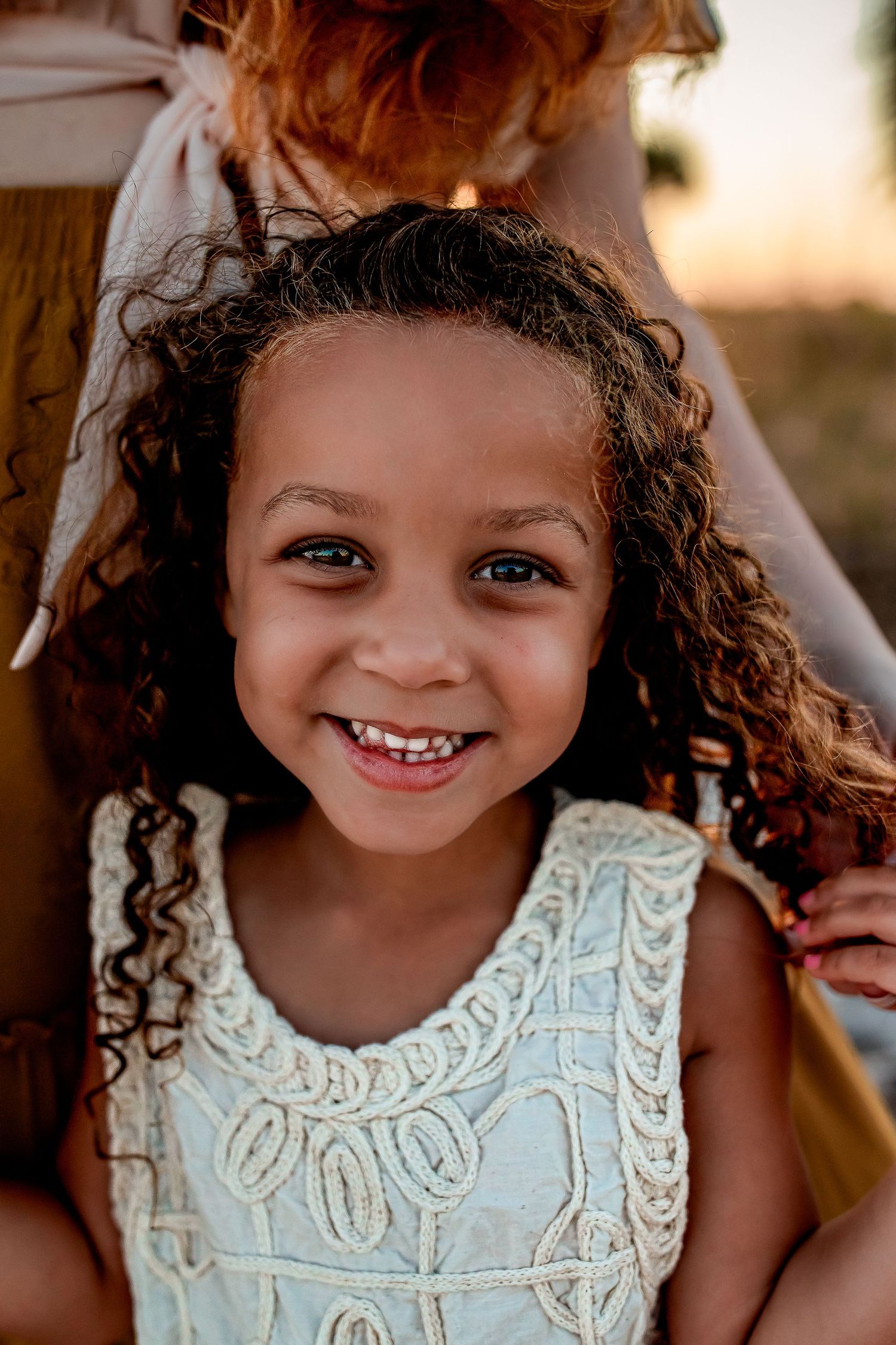 Adorable curly-haired little girl in white lace dress smiles brightly at golden hour.