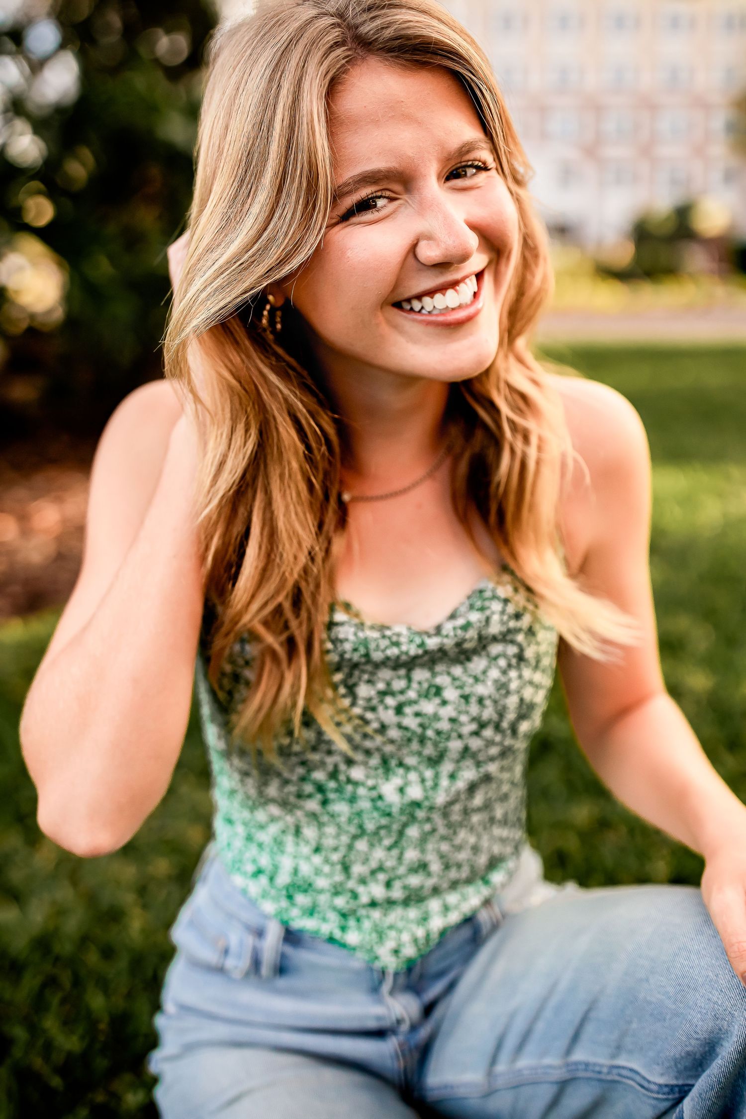 Cheerful blonde woman in green sequin top laughs while sitting on grass in sunny outdoor setting.