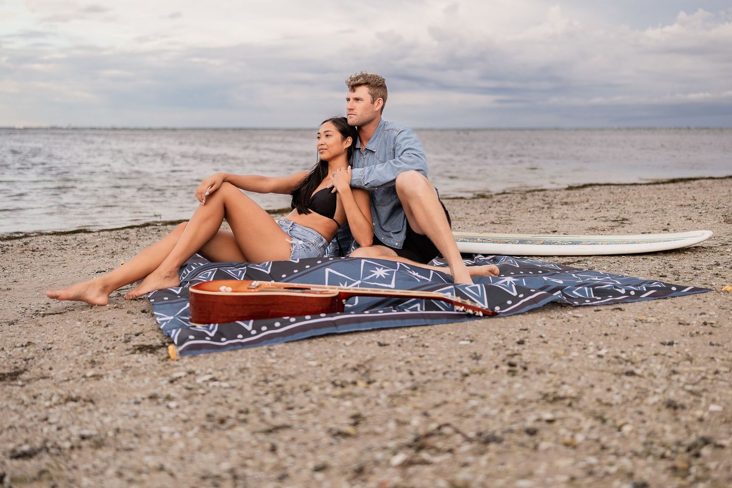 Couple relaxing on a beach blanket near a surfboard with calm ocean views behind them.
