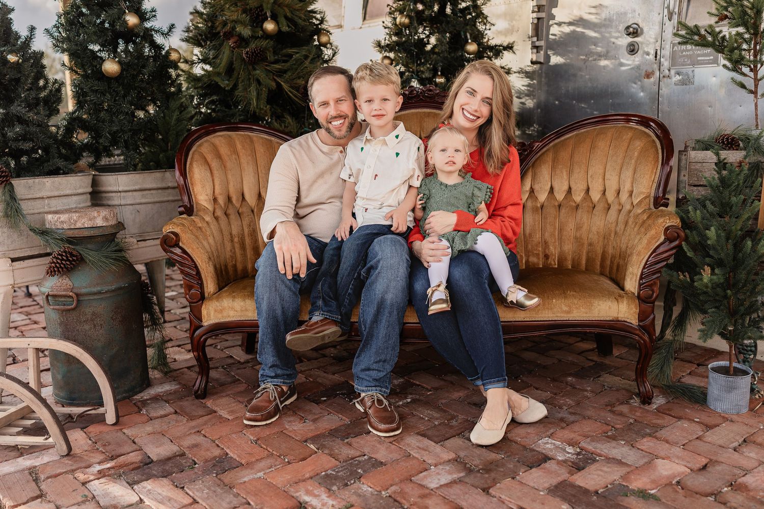 A family of four sits on a vintage sofa surrounded by Christmas trees on a brick patio.