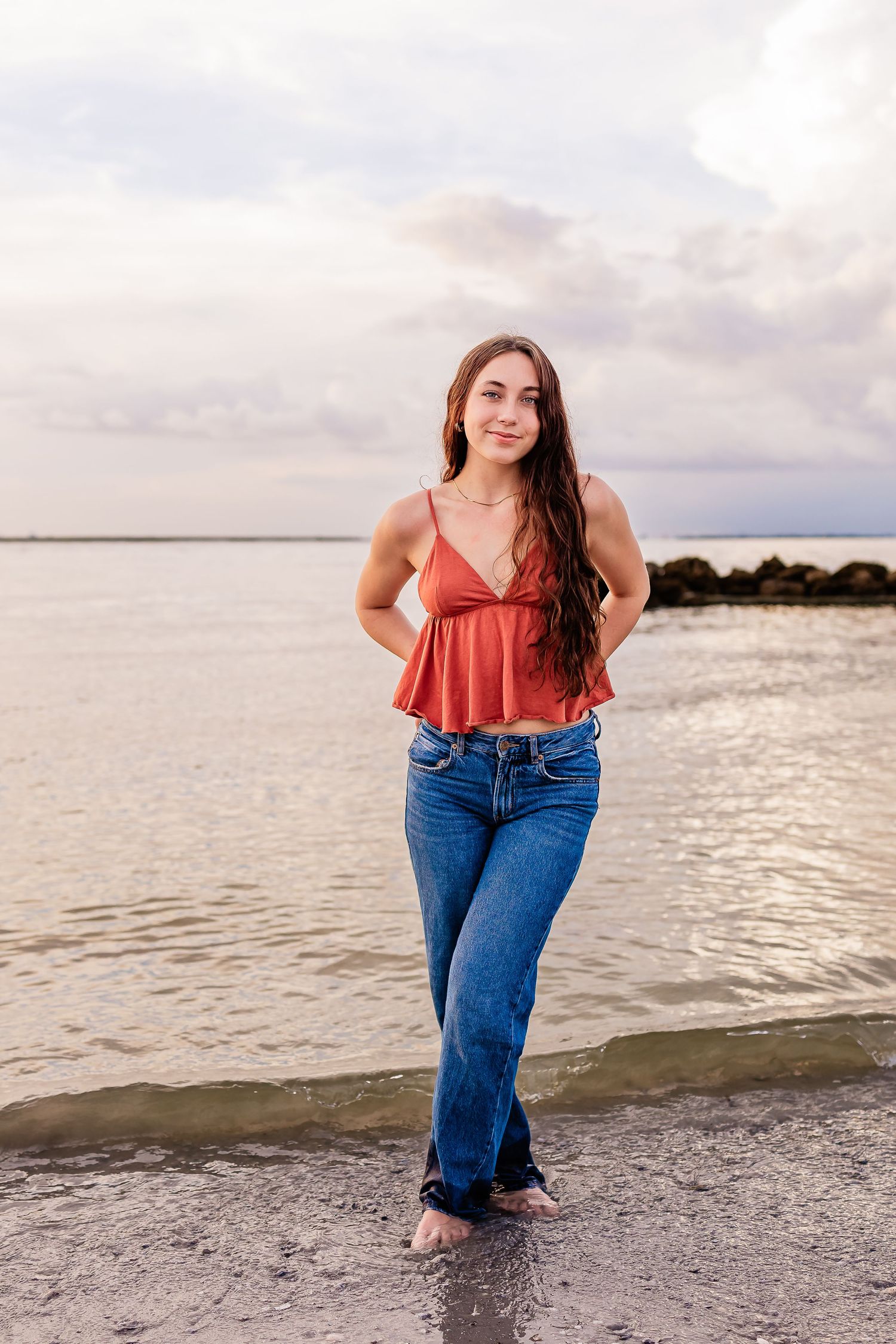 Young woman in red crop top and flare jeans standing barefoot on a rocky shoreline at sunset.
