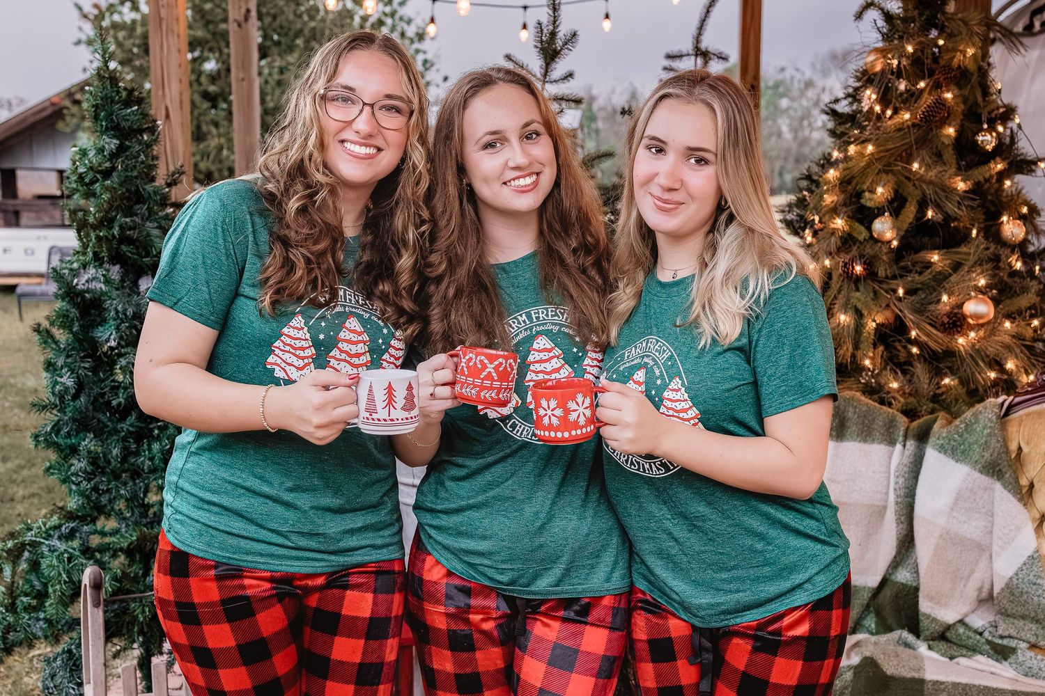 High school senior friends in matching green Christmas shirts and red plaid pajamas holding holiday mugs near a decorated tree.
