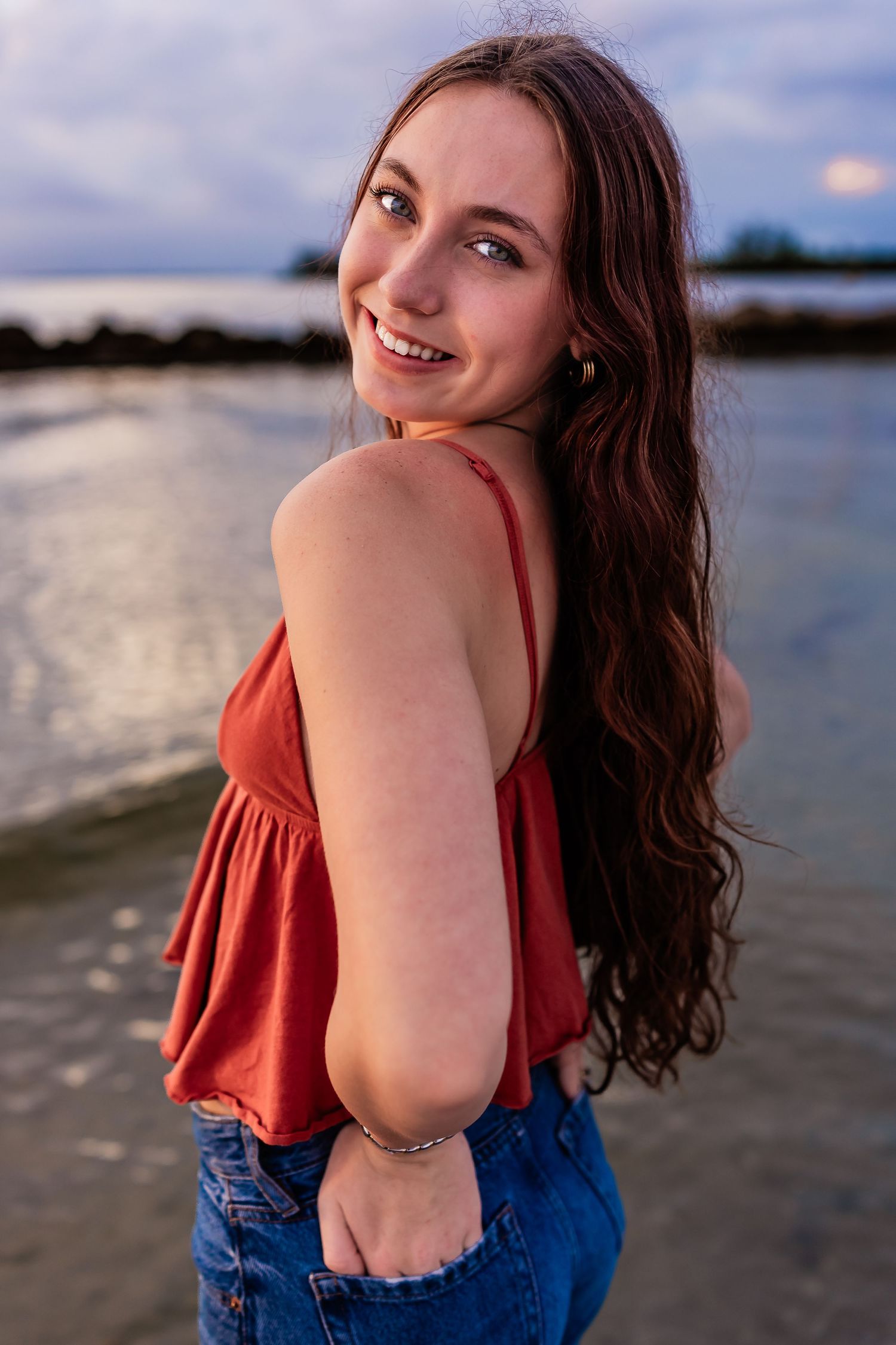 Young woman with long brown hair in red top and jeans poses near water at dusk.