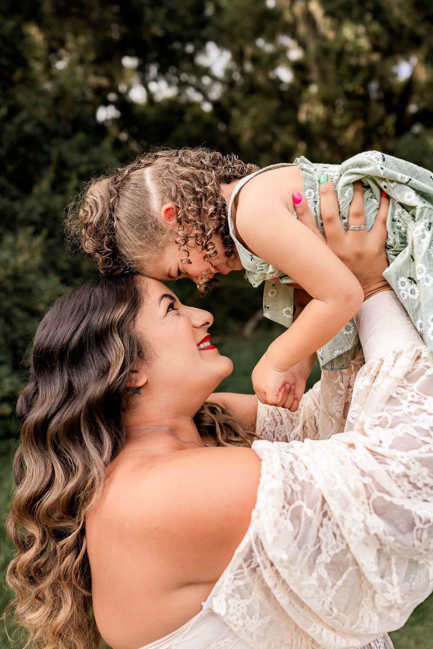 A joyful mother in white lace dress lifting her curly-haired toddler in a green outfit outdoors.