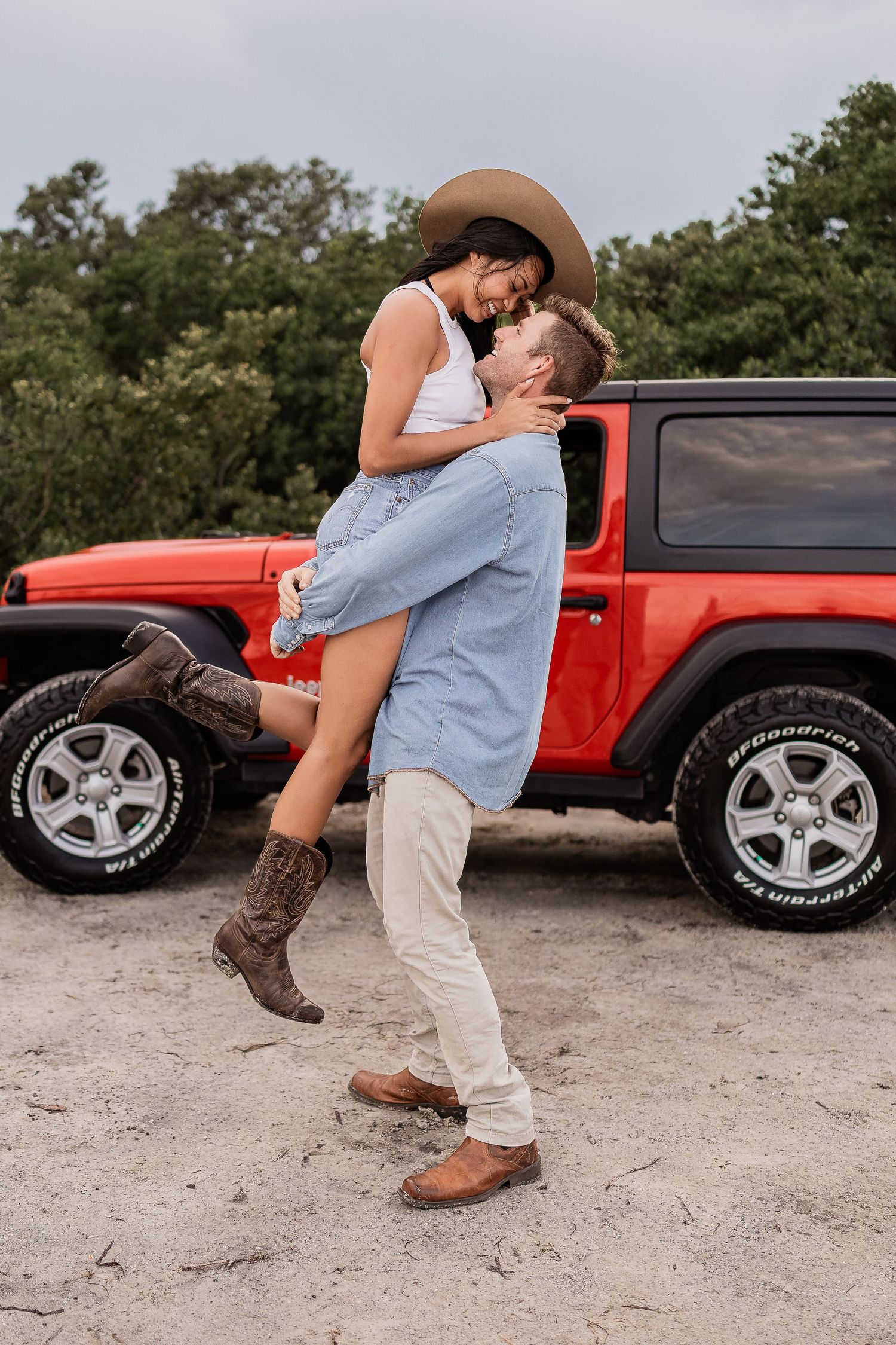 Man lifting and spinning woman in hat near red Jeep on sandy terrain.