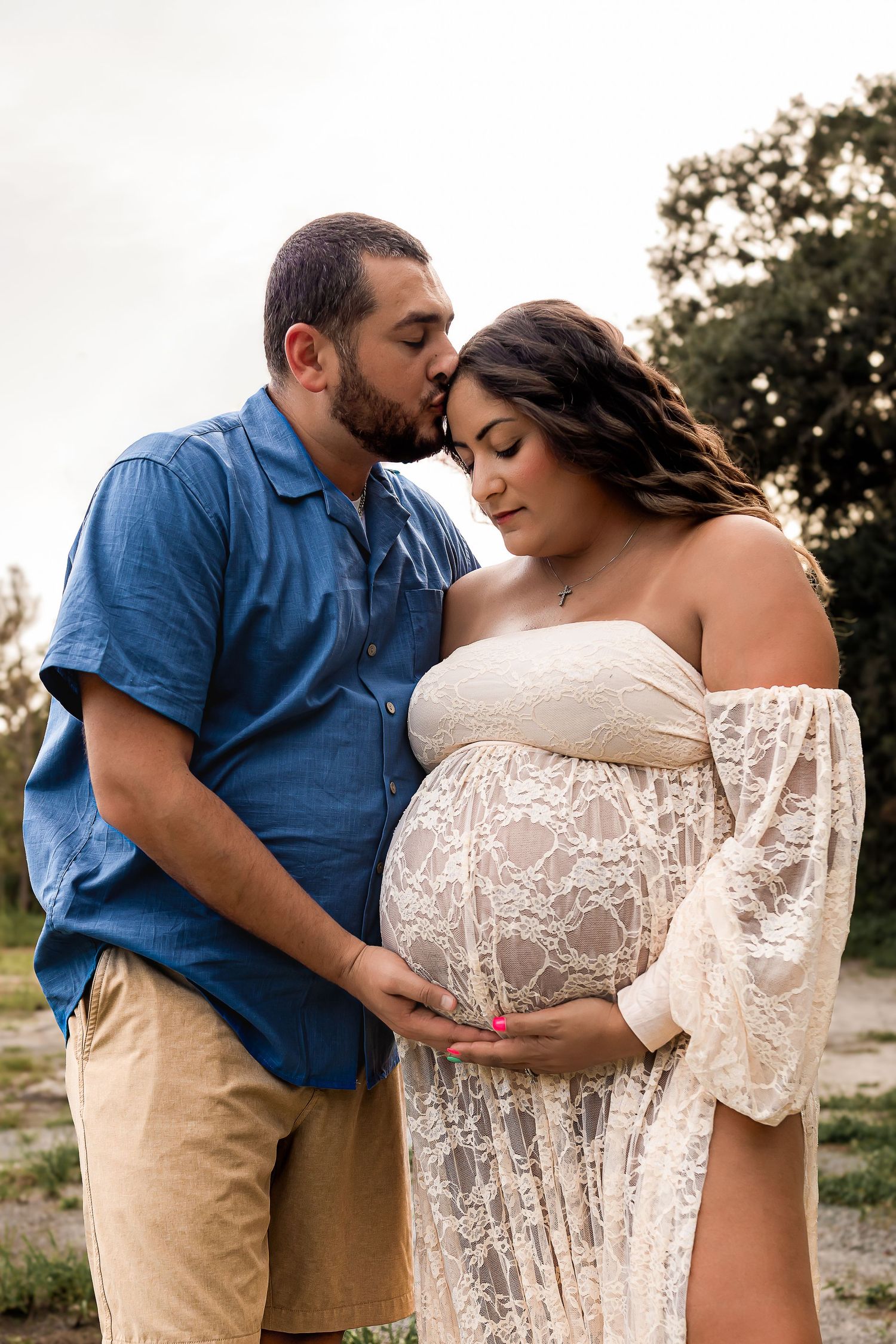 A tender moment as a man in blue shirt kisses his pregnant partner in a white lace gown outdoors.