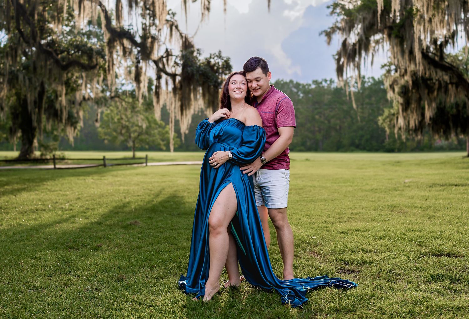 Couple poses in green park, woman in flowing blue maternity gown embraced by man in pink shirt.