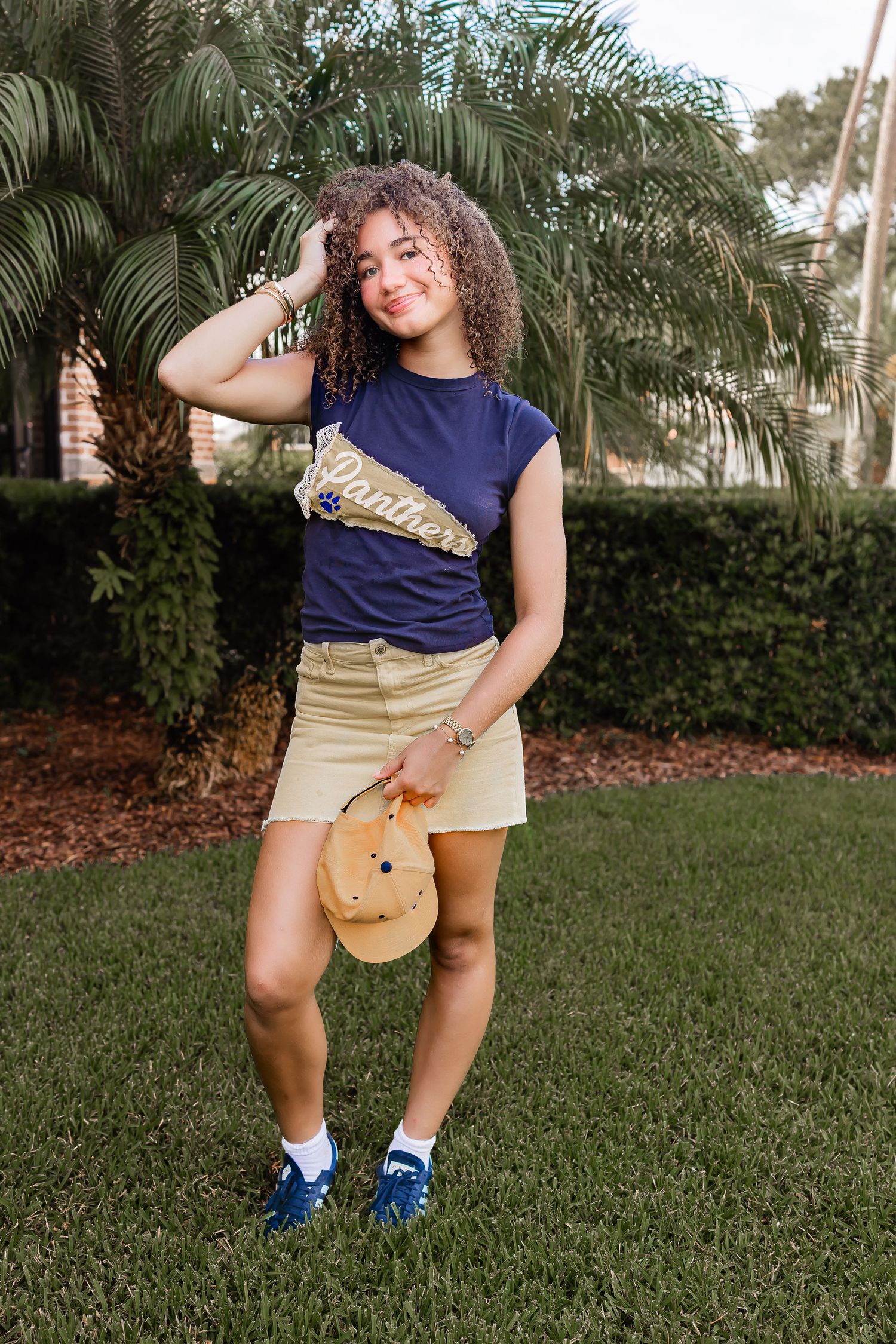 Young womanl in Panthers tee and khaki skirt holding hat near palm trees.