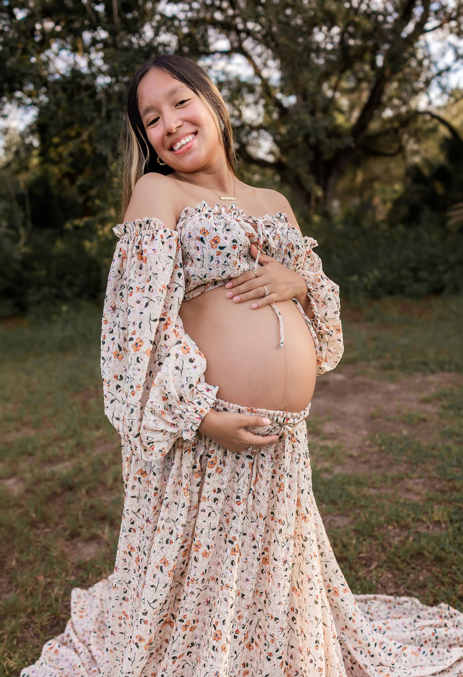 Pregnant woman in floral off-shoulder dress cradling baby bump outdoors.