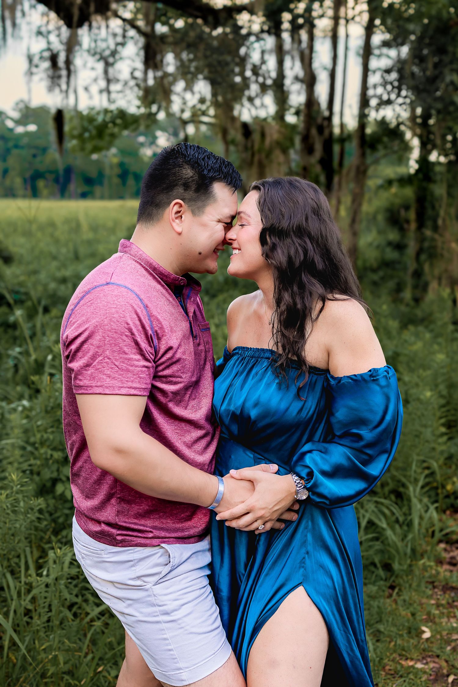 Couple nearly kissing in lush green field, woman in elegant blue off-shoulder maternity dress.