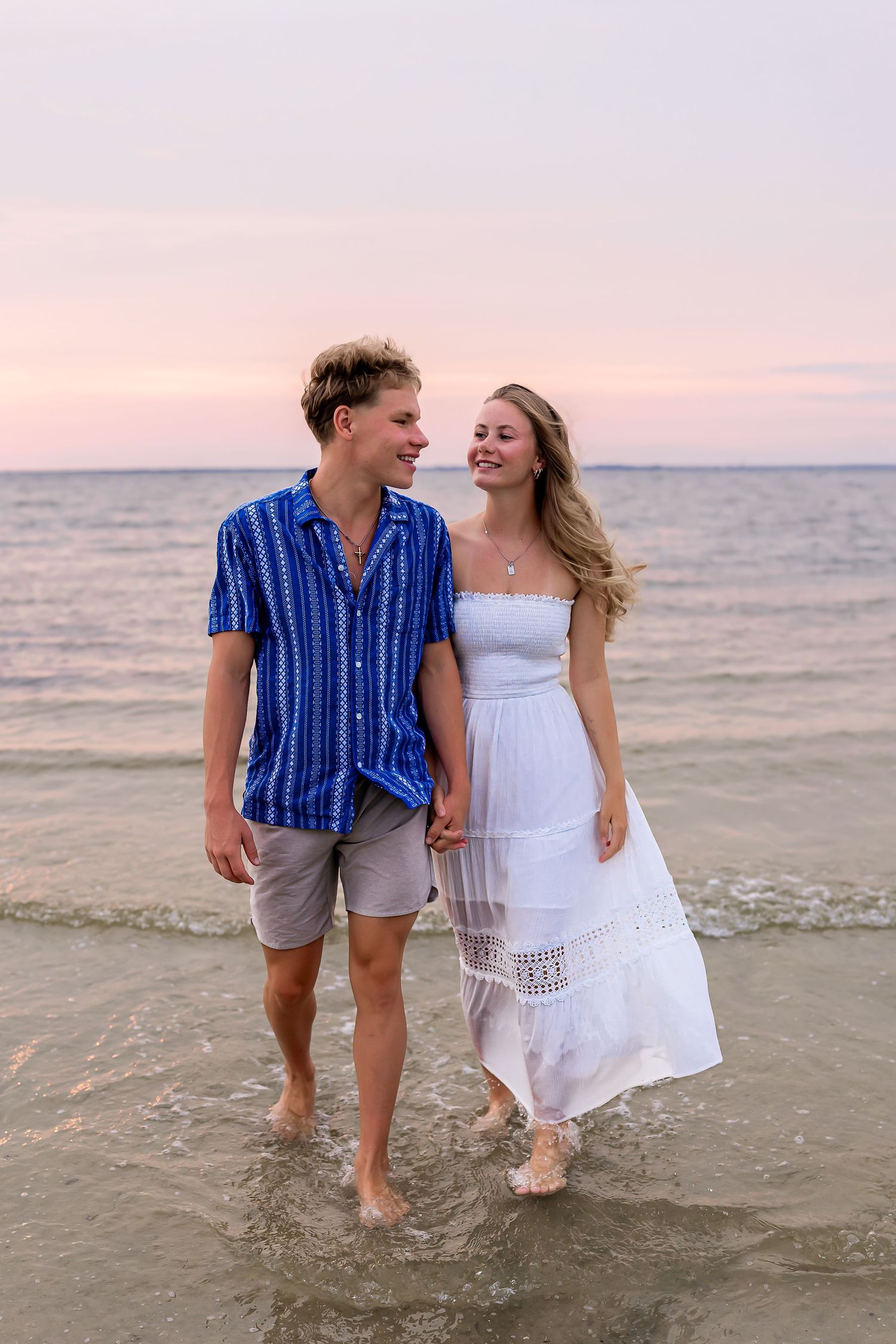 Young couple walking hand in hand on beach at sunset, woman in white dress, man in blue shirt.