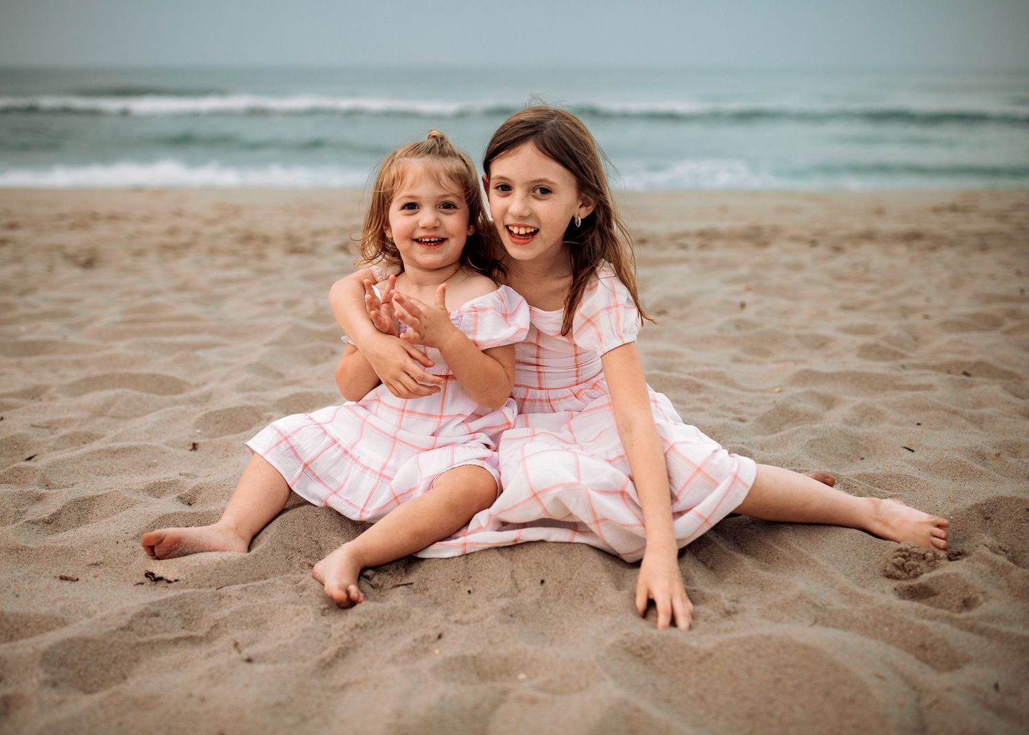 Two young sisters in matching pink plaid dresses sit together smiling on a sandy beach.